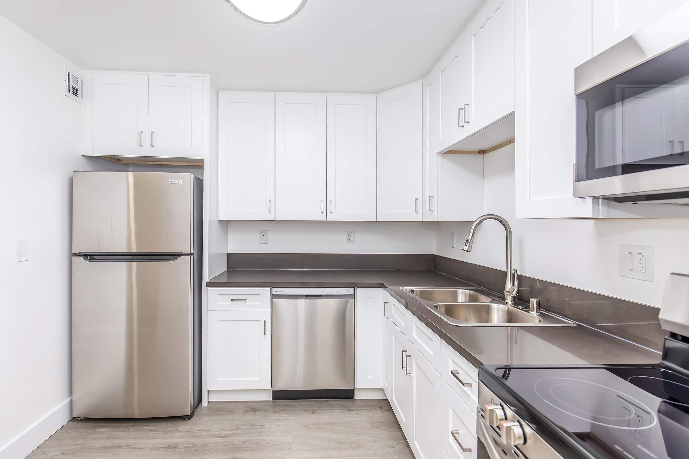 Modern kitchen featuring white cabinetry, stainless steel appliances including a refrigerator and dishwasher, and a sleek countertop. The space is well-lit with an overhead light and has a clean, minimalist design, ideal for cooking and entertaining.