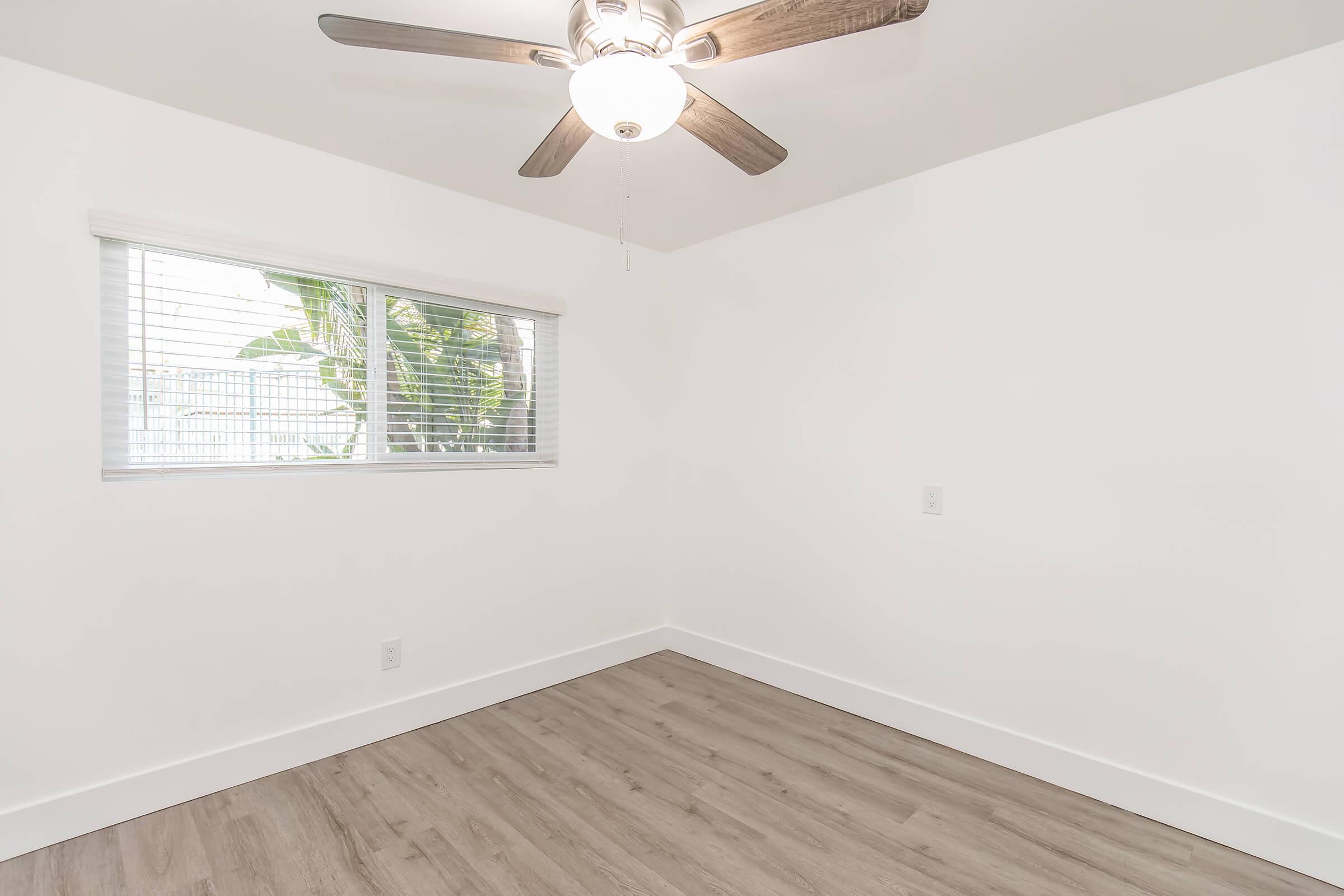 A minimalist room featuring light-colored walls, a window with blinds allowing natural light, and wooden flooring. The ceiling has a modern ceiling fan with five blades. The space is empty, showcasing a clean and simple design, perfect for various uses.