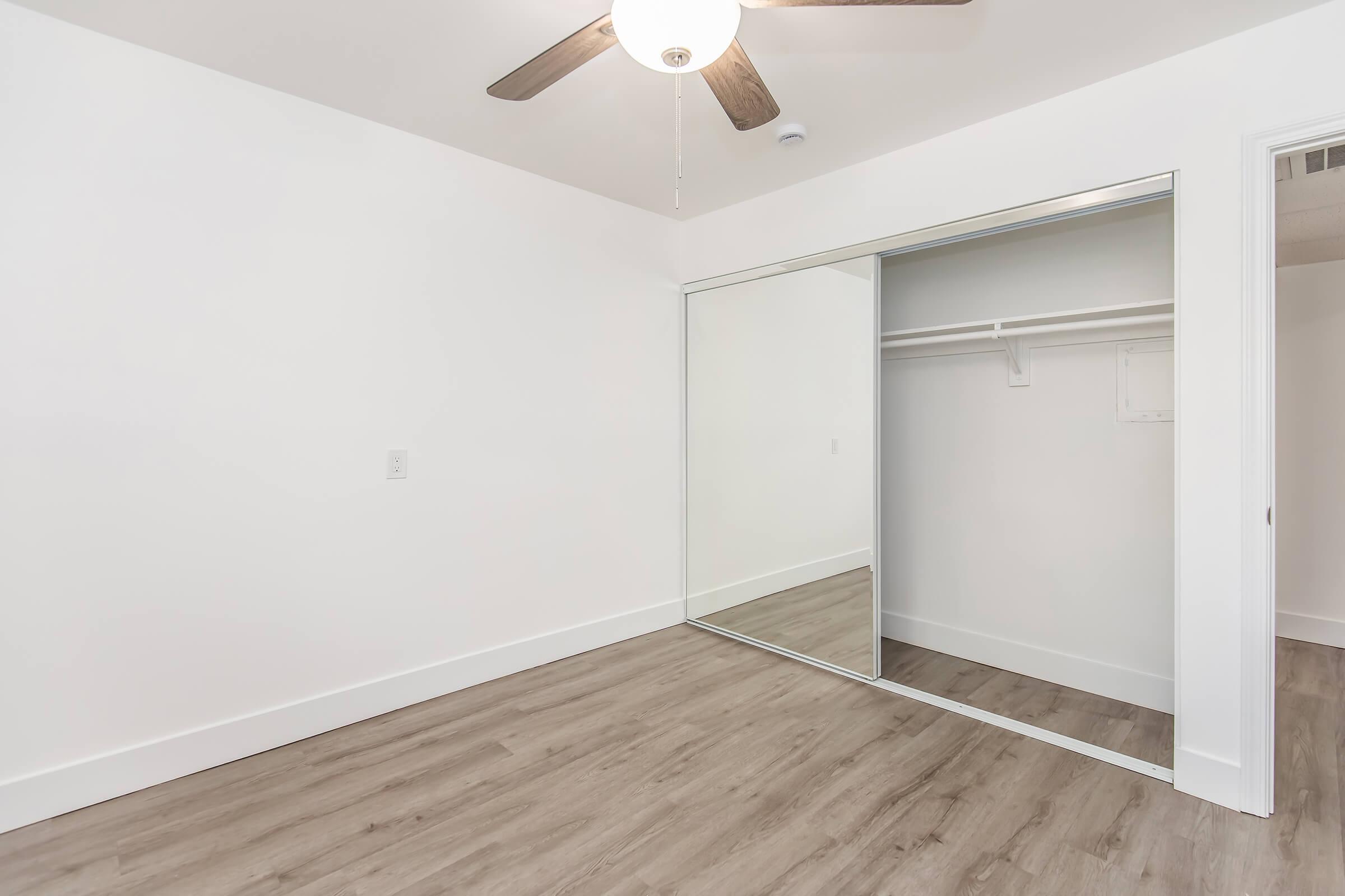 Empty room with light-colored walls and wooden flooring. A ceiling fan with wooden blades is visible. There’s a mirrored wardrobe with sliding doors on one side, reflecting the room's interior. The space appears bright and well-lit.