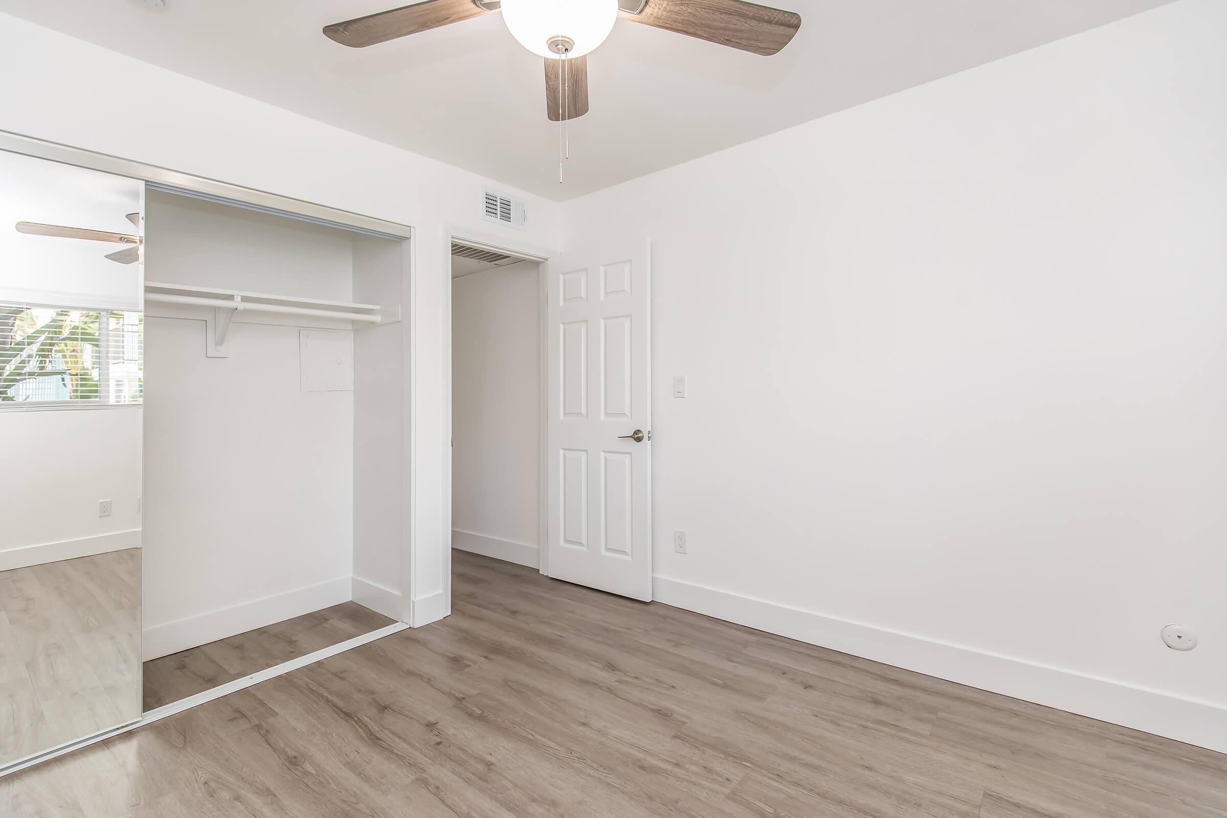 Empty room with light-colored walls and wooden flooring. There is a ceiling fan and a mirrored closet door on one side. A door leads to another area, and a window can be seen in the background, allowing natural light to enter. The space is clean and well-lit, suitable for various uses.