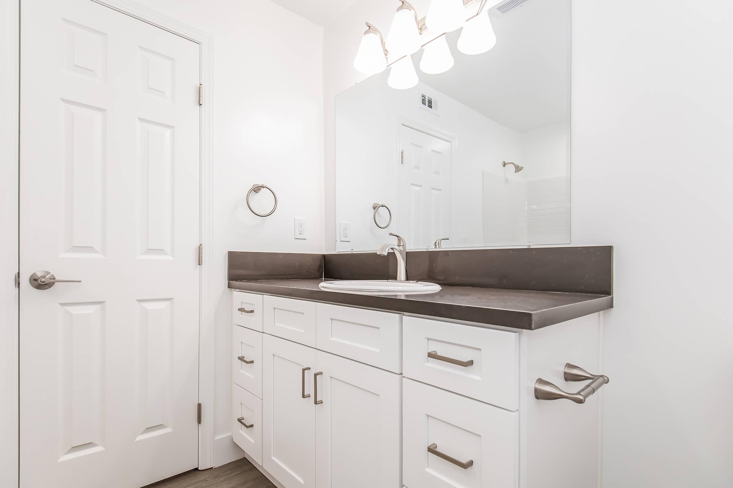 A modern bathroom featuring a white vanity with a dark countertop, a round mirror above, and a polished faucet. The walls are painted white, and there's a towel rack and door visible. Bright lighting is provided by three fixtures above the mirror.