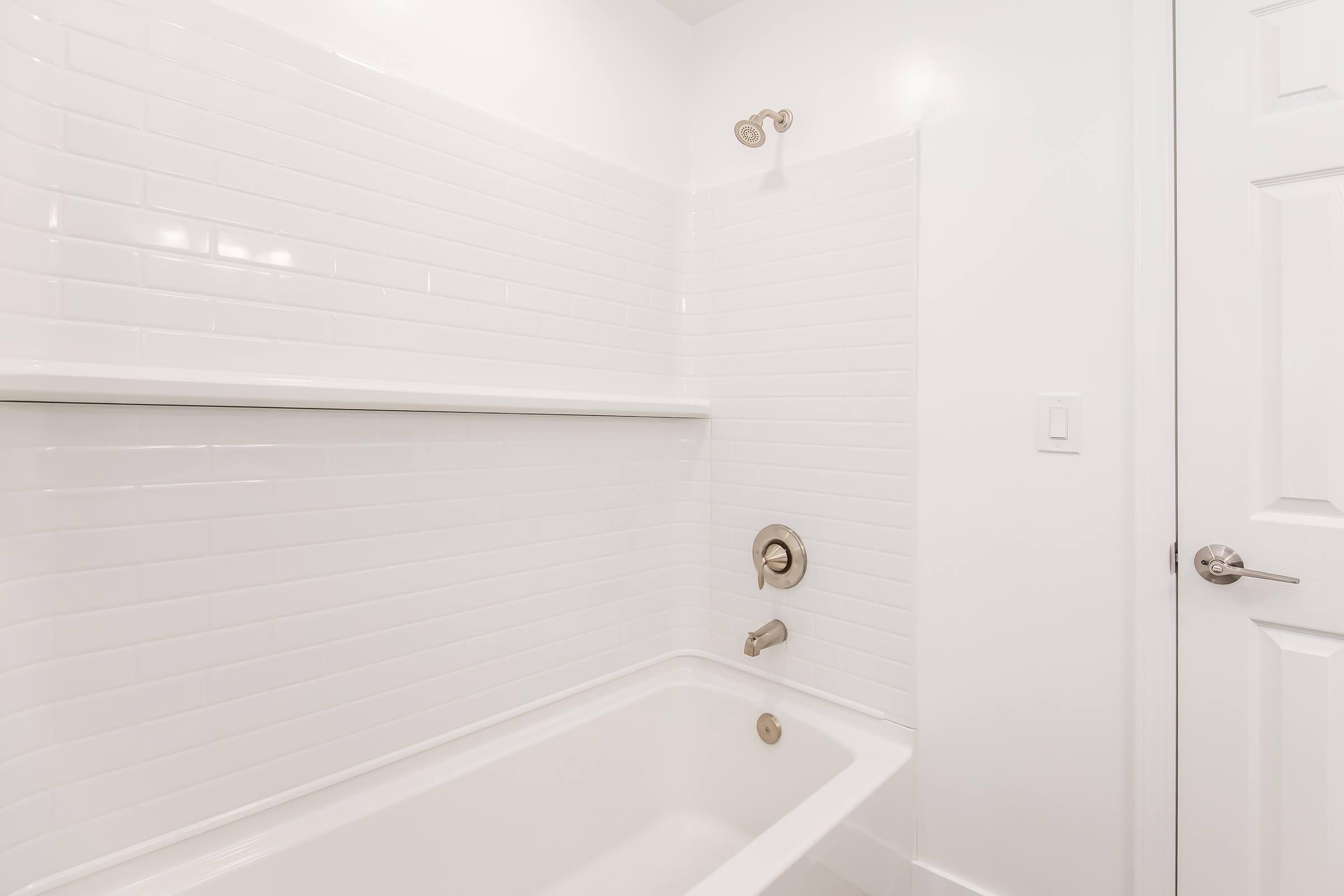 A clean, modern bathroom featuring white tiled walls and a bathtub with an integrated shower. The space includes a sleek faucet and showerhead, along with a shelf above the tub for storage. A white door is visible, contributing to the bright, minimalist design.