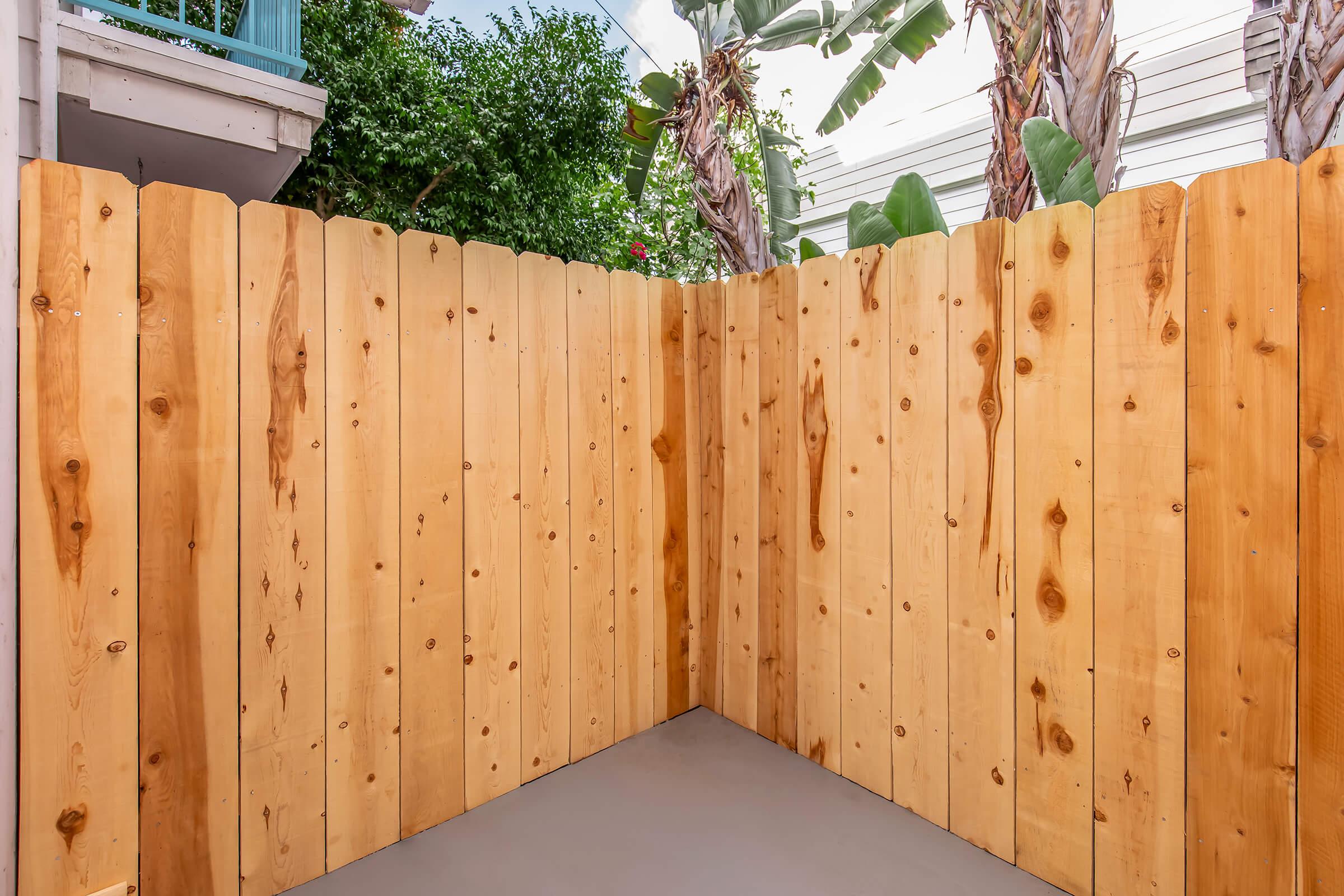 A wooden fence made of light-colored planks forms a corner in an outdoor space. The fence is topped with a slightly curved, scalloped edge and features various knots and grains. In the background, lush green plants and palm trees are visible, creating a tropical atmosphere.