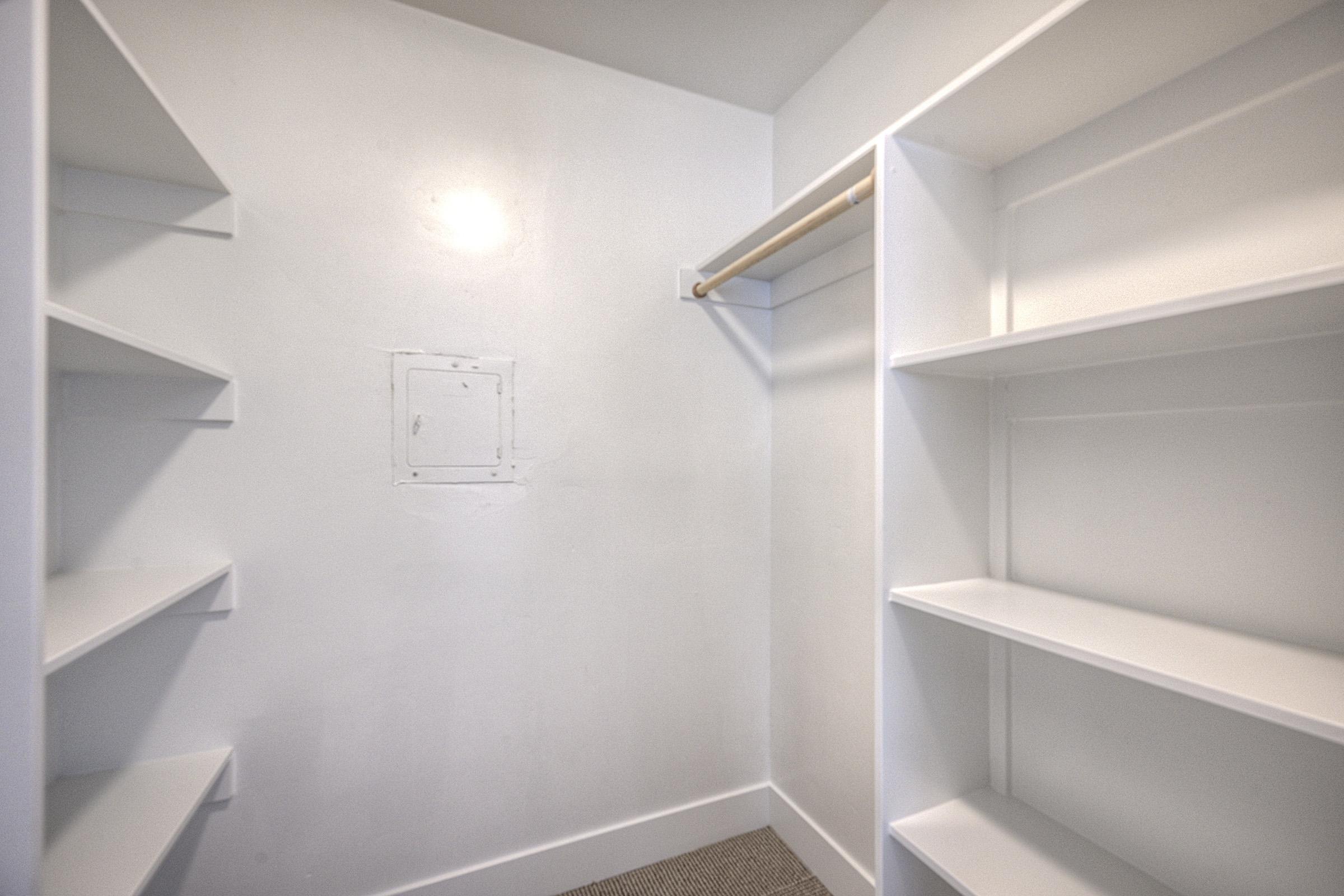 A clean, empty closet featuring white walls and shelving units on either side. The space includes a simple wooden rod for hanging clothes, with light colored carpeting on the floor. The overall appearance is bright and minimalistic, ideal for storage or organization.