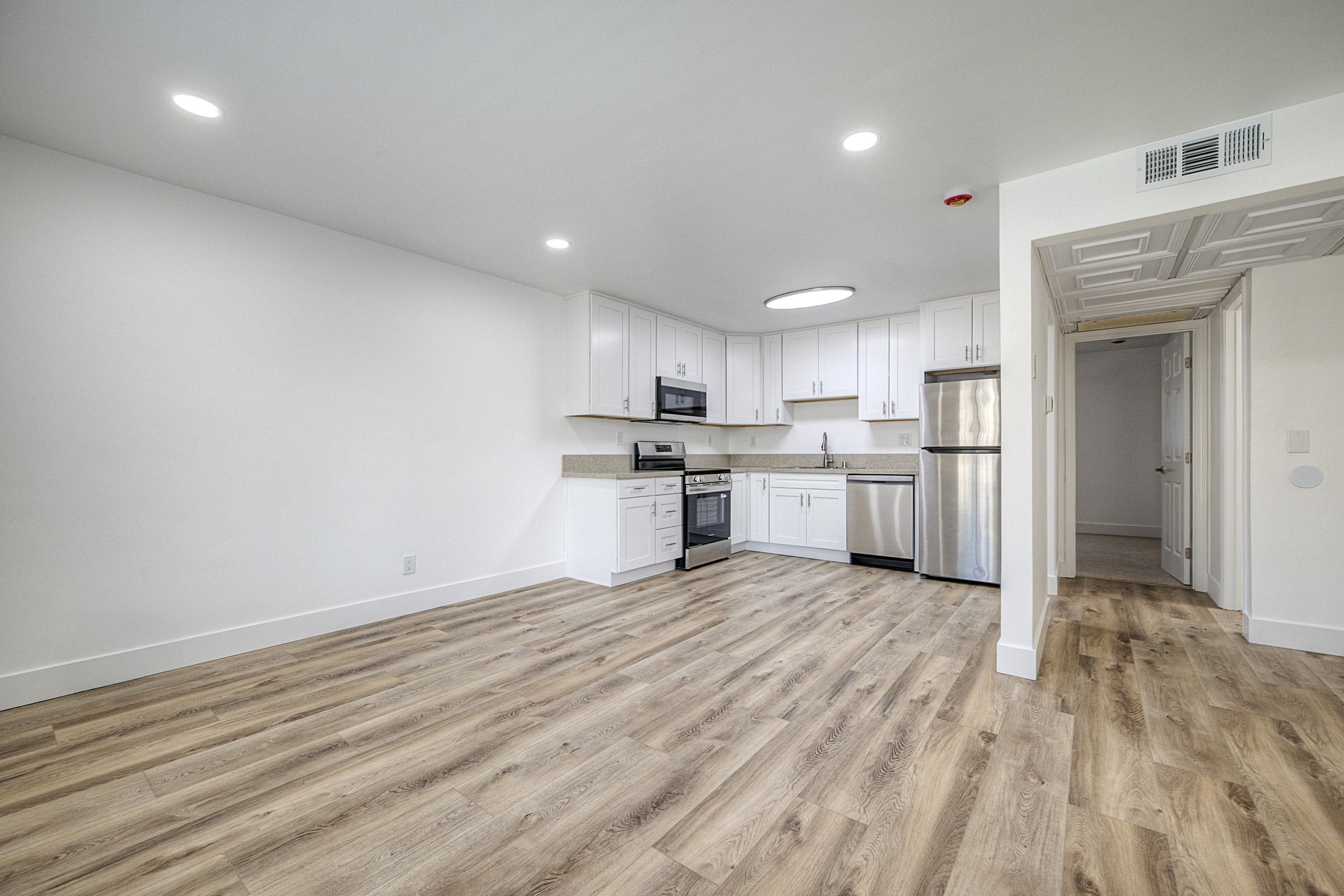 A spacious, modern kitchen with white cabinets, stainless steel appliances, and wood-look flooring. The layout includes a microwave, stove, refrigerator, and a small island. Bright overhead lighting illuminates the room, which is connected to a hallway leading to another area. The walls are painted white, enhancing the open feel.