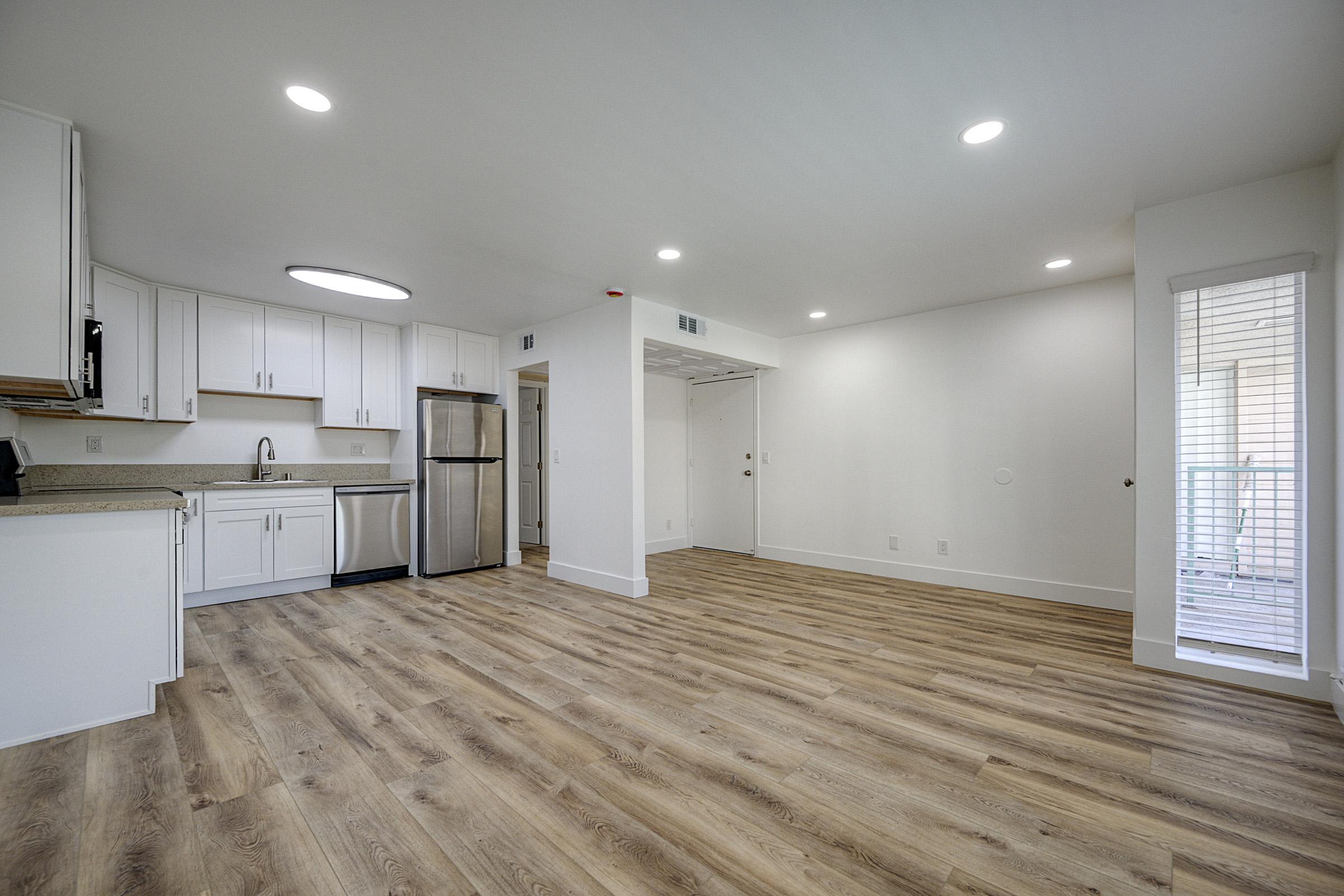 A modern, empty living space featuring a kitchen with white cabinets, a stainless steel refrigerator, and a countertop. The floor is light-colored wood laminate. The area has overhead lighting and a doorway leading outside. The walls are painted white, creating a bright and airy atmosphere.