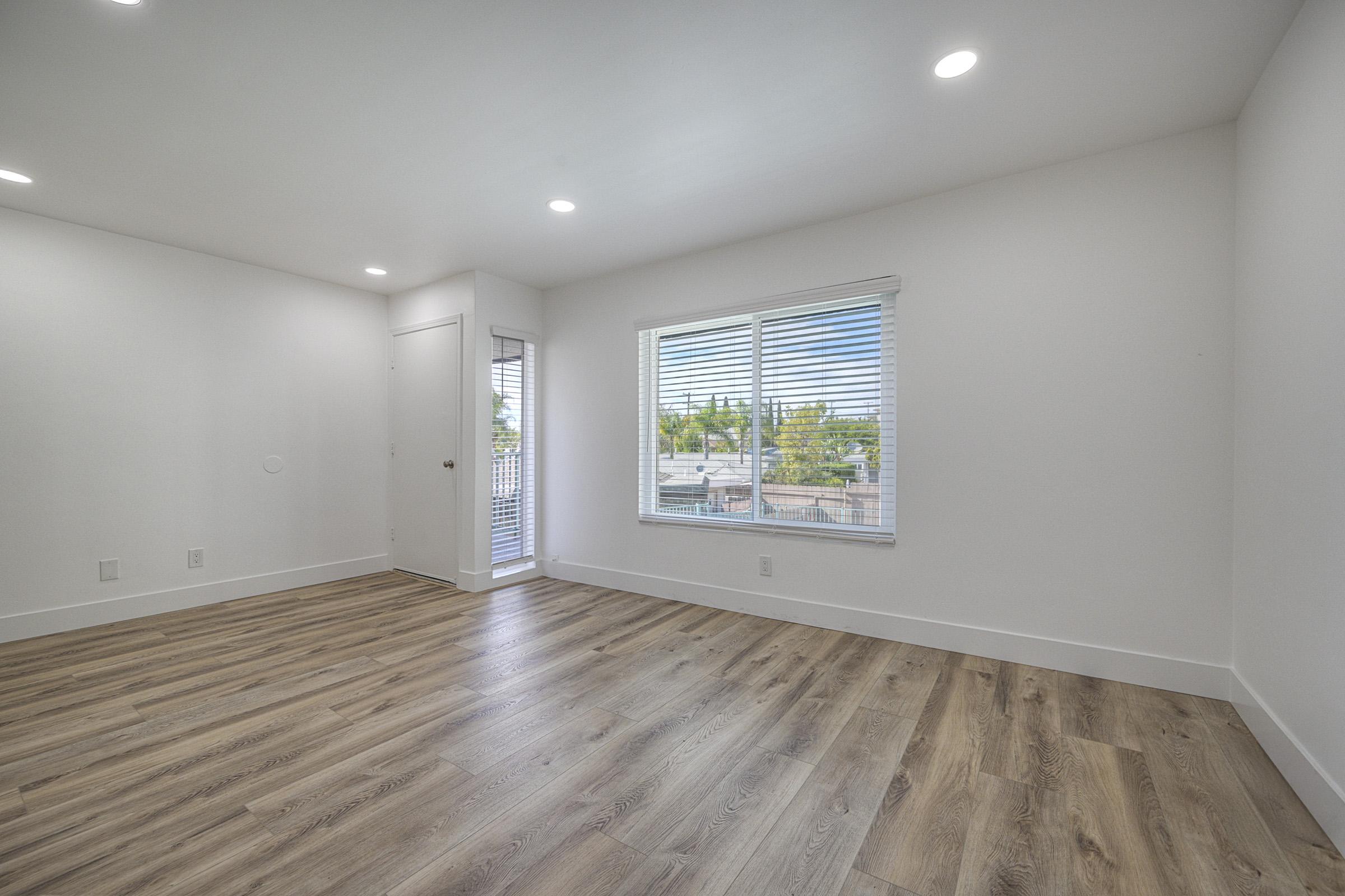Empty room with light-colored walls and wooden flooring. A window with blinds lets in natural light, and there's a door visible on the left side. The space is minimalistic, featuring recessed lighting and no furnishings.