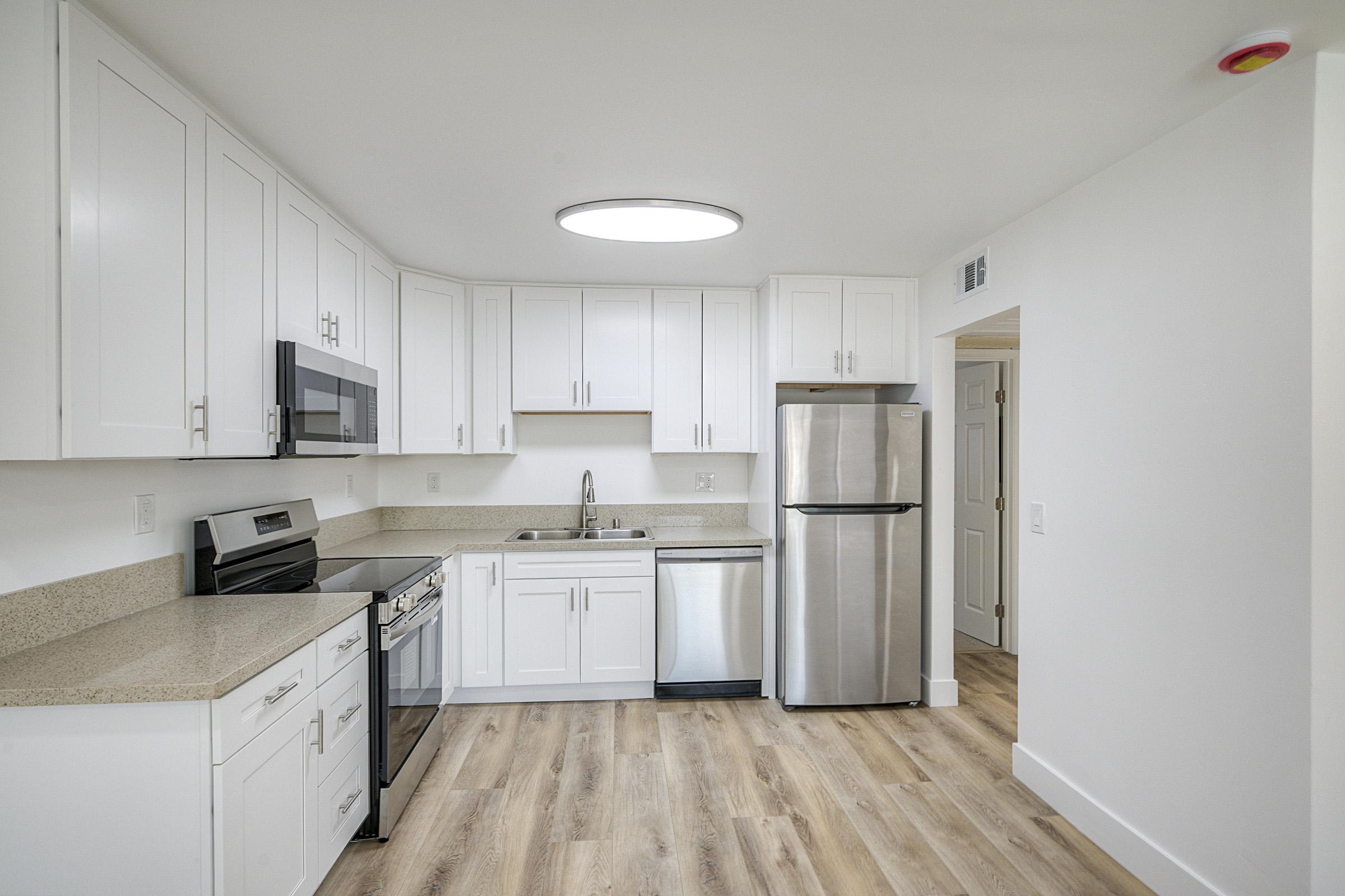 A modern kitchen featuring white cabinets, a stainless steel fridge, a microwave, and an oven. The countertop is light-colored with a subtle pattern. There is a sink in the center, and large windows provide natural lighting. The flooring is light wood, enhancing the contemporary feel of the space.