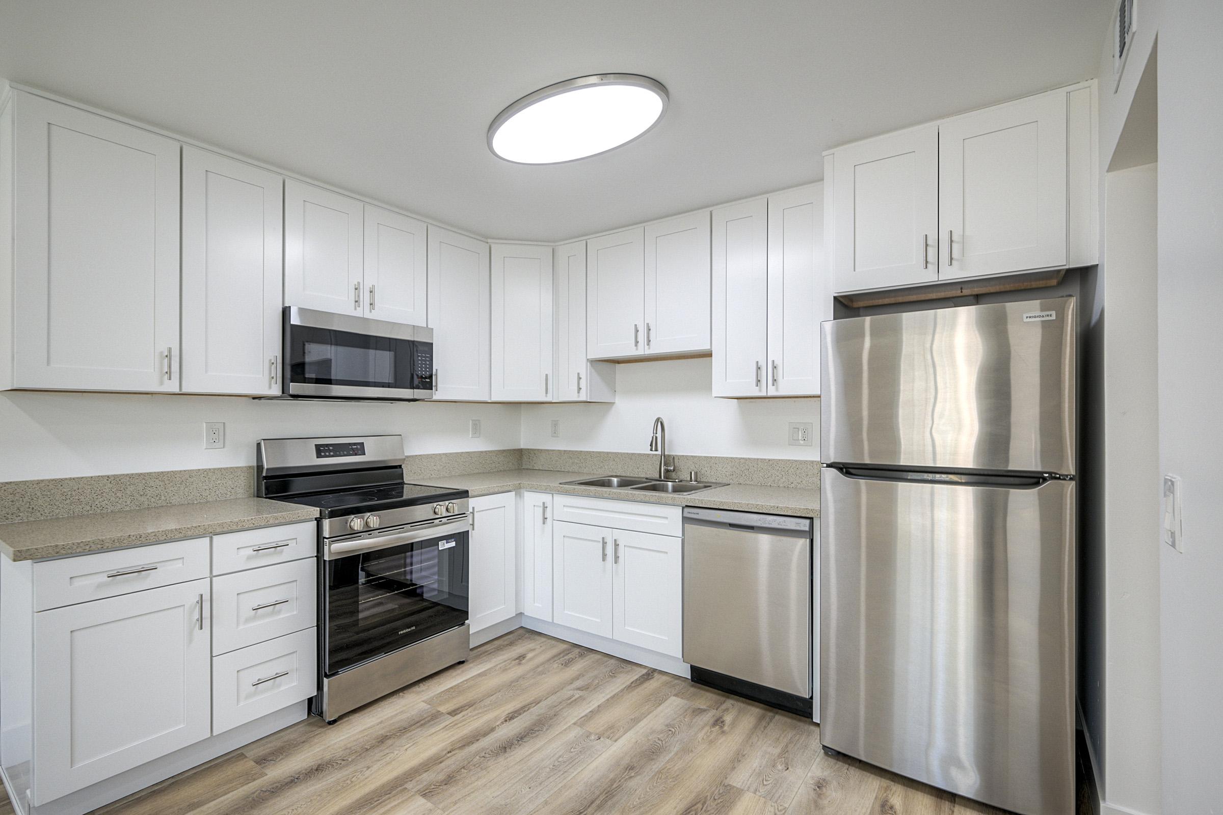 Modern kitchen featuring white cabinetry, stainless steel appliances including an oven, microwave, and refrigerator. The countertops are a light color, and the flooring is a light wood finish. The room is well-lit with a circular ceiling light.