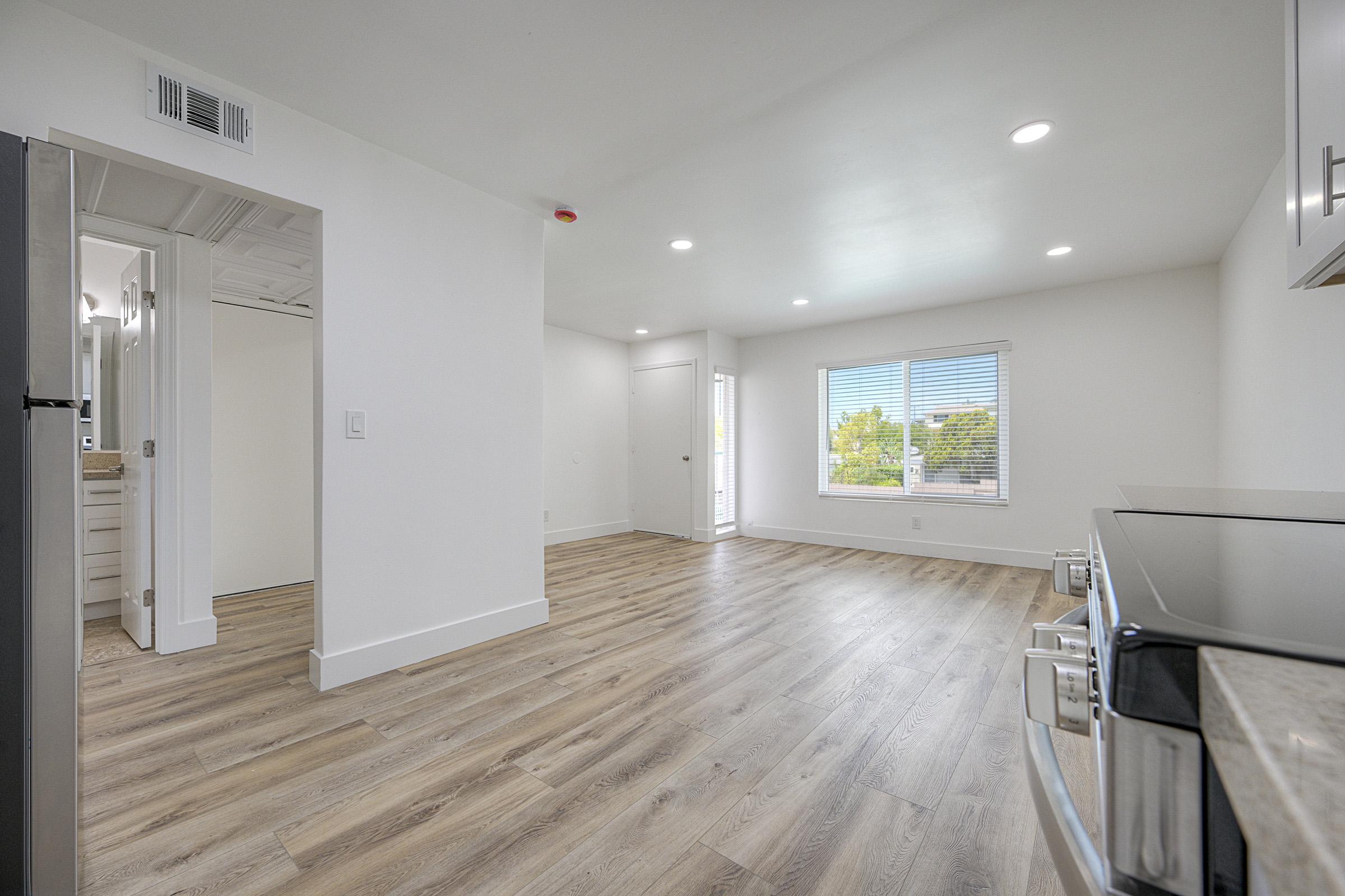 Bright, modern living space featuring light hardwood flooring, white walls, and recessed lighting. The room has a large window with blinds, allowing natural light. A glimpse of a bathroom is visible through an open door, and there is a kitchen area with a stainless steel microwave.
