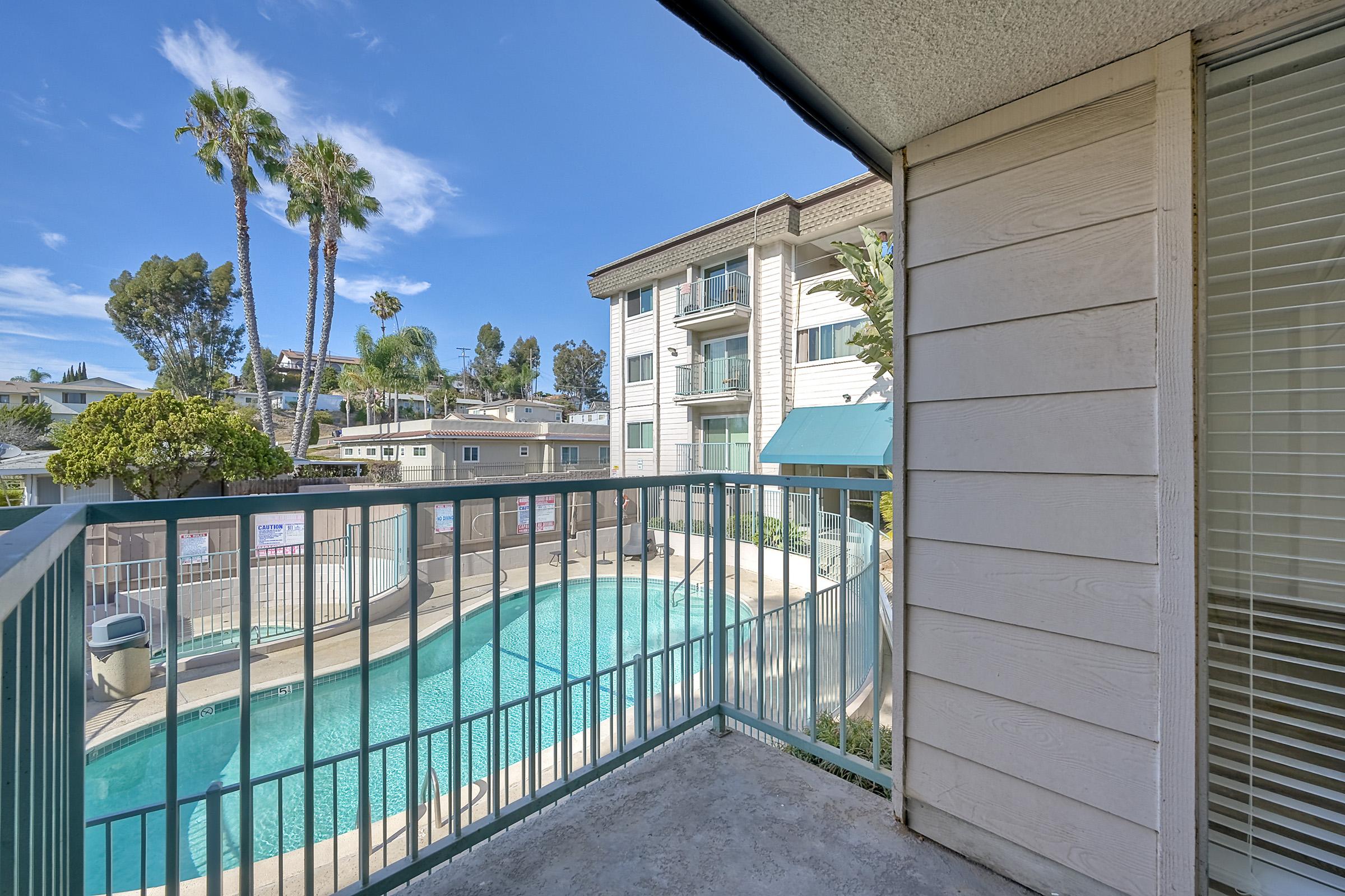 View from a balcony overlooking a pool area surrounded by palm trees and residential buildings. The sky is clear with a few clouds, and sunlight is casting shadows on the balcony railing. The scene reflects a serene and inviting atmosphere typical of a warm climate.