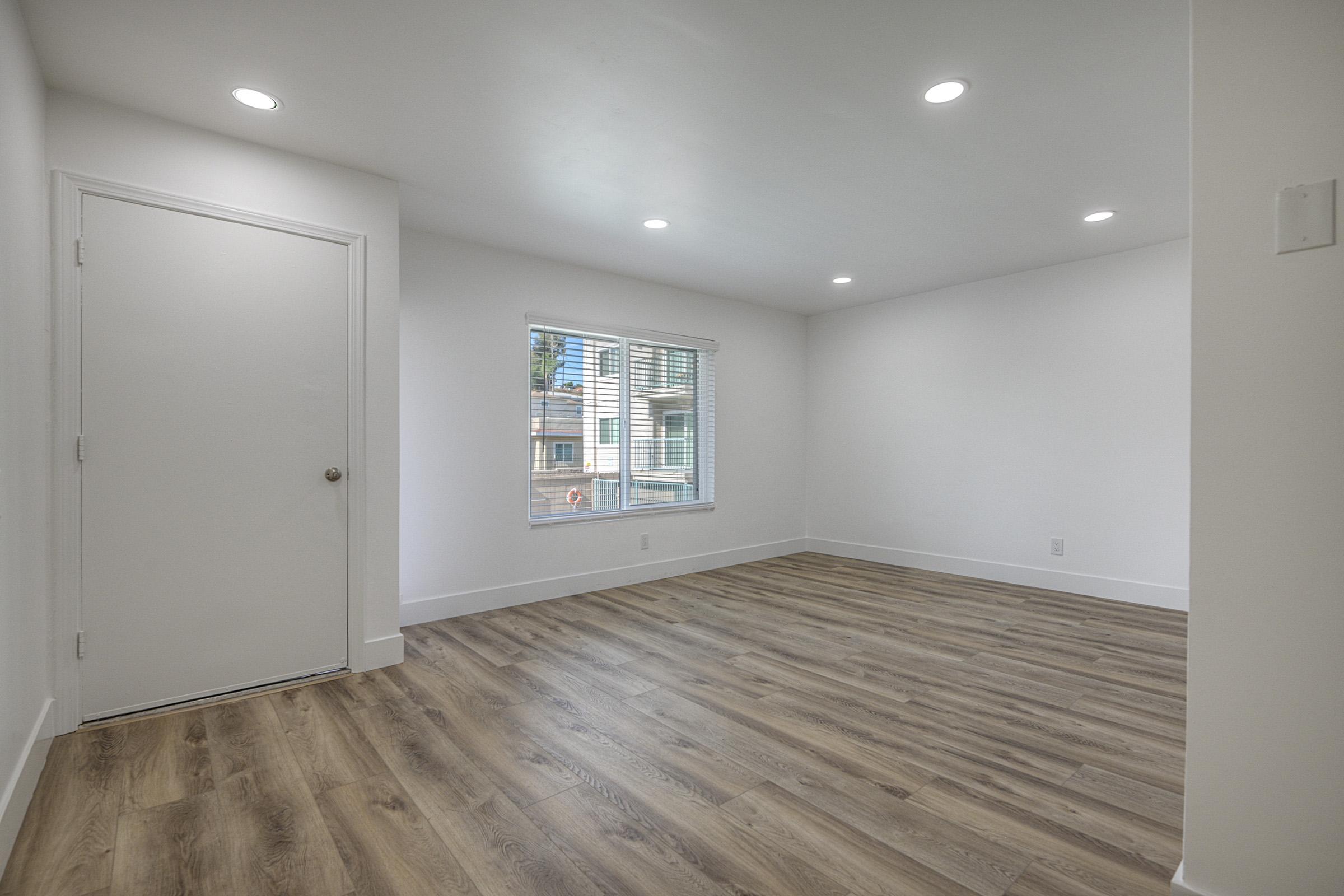A spacious, well-lit empty room with white walls, wooden flooring, and recessed lighting. There is a single window with blinds allowing natural light, and a door on the left side leading to another space. The overall atmosphere is bright and ready for decoration.