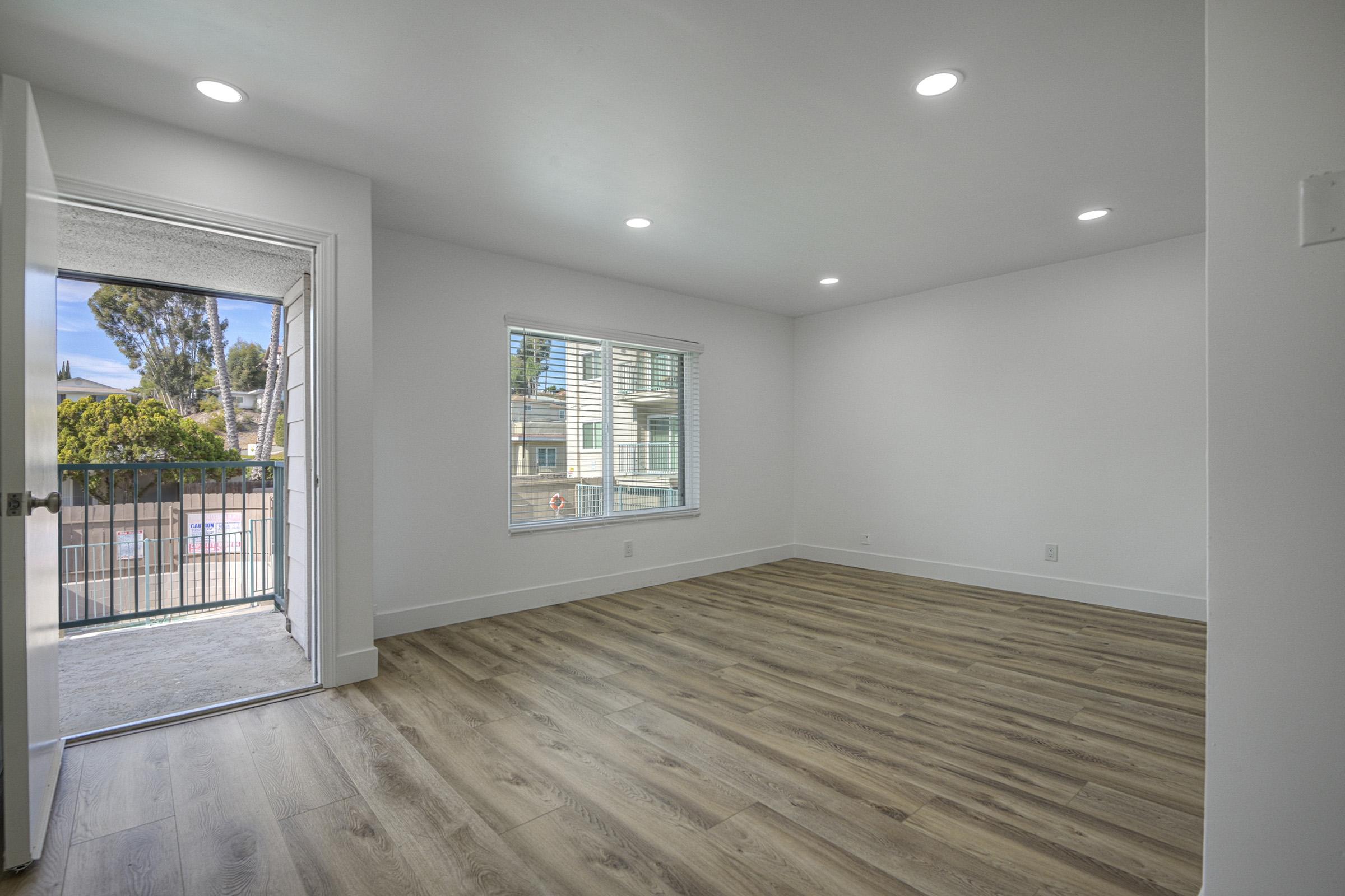 A bright, empty living room with light-colored walls and wooden flooring. A window and a door leading to a balcony are visible, allowing natural light to fill the space. The room has recessed lighting, creating a modern and spacious feel. Ideal for residential use.