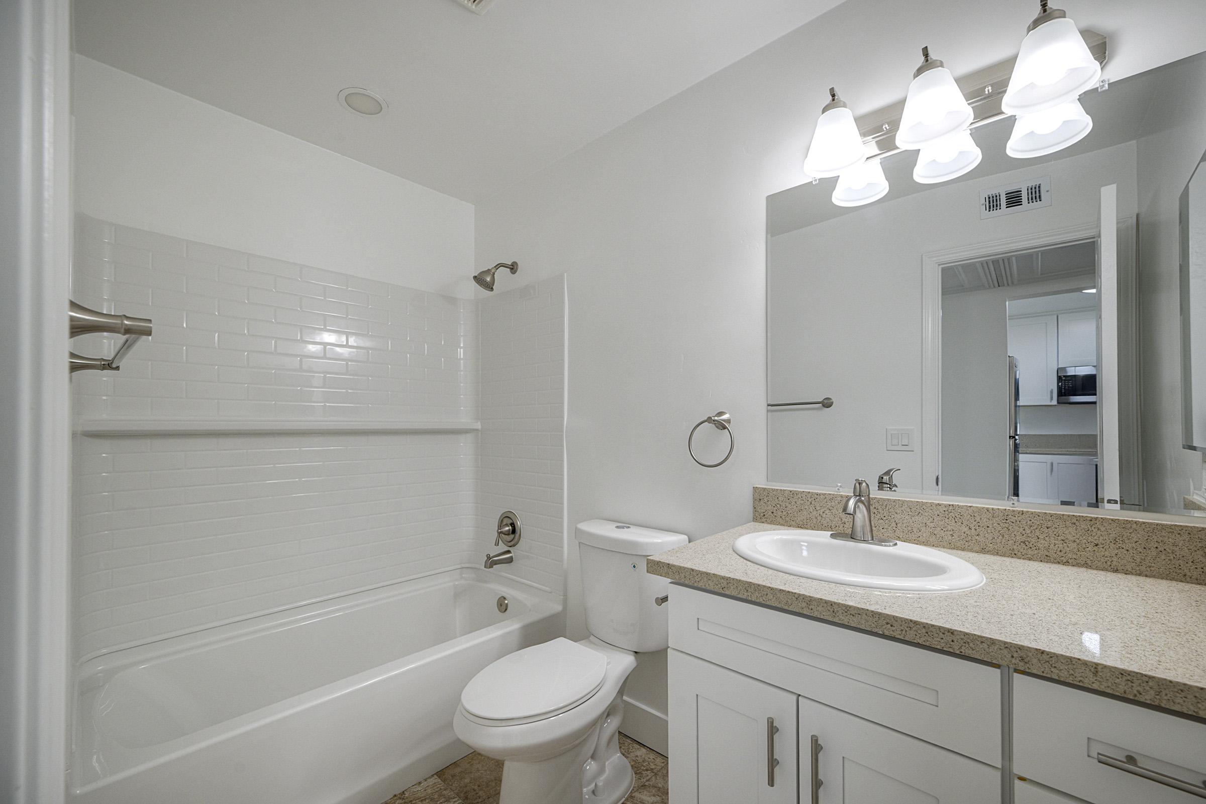 A modern bathroom featuring a white tiled bathtub and shower combination, light granite countertop with a sink, a wall-mounted mirror, and a lighting fixture with three bulbs. The walls are painted white, and there is a towel holder beside the sink.