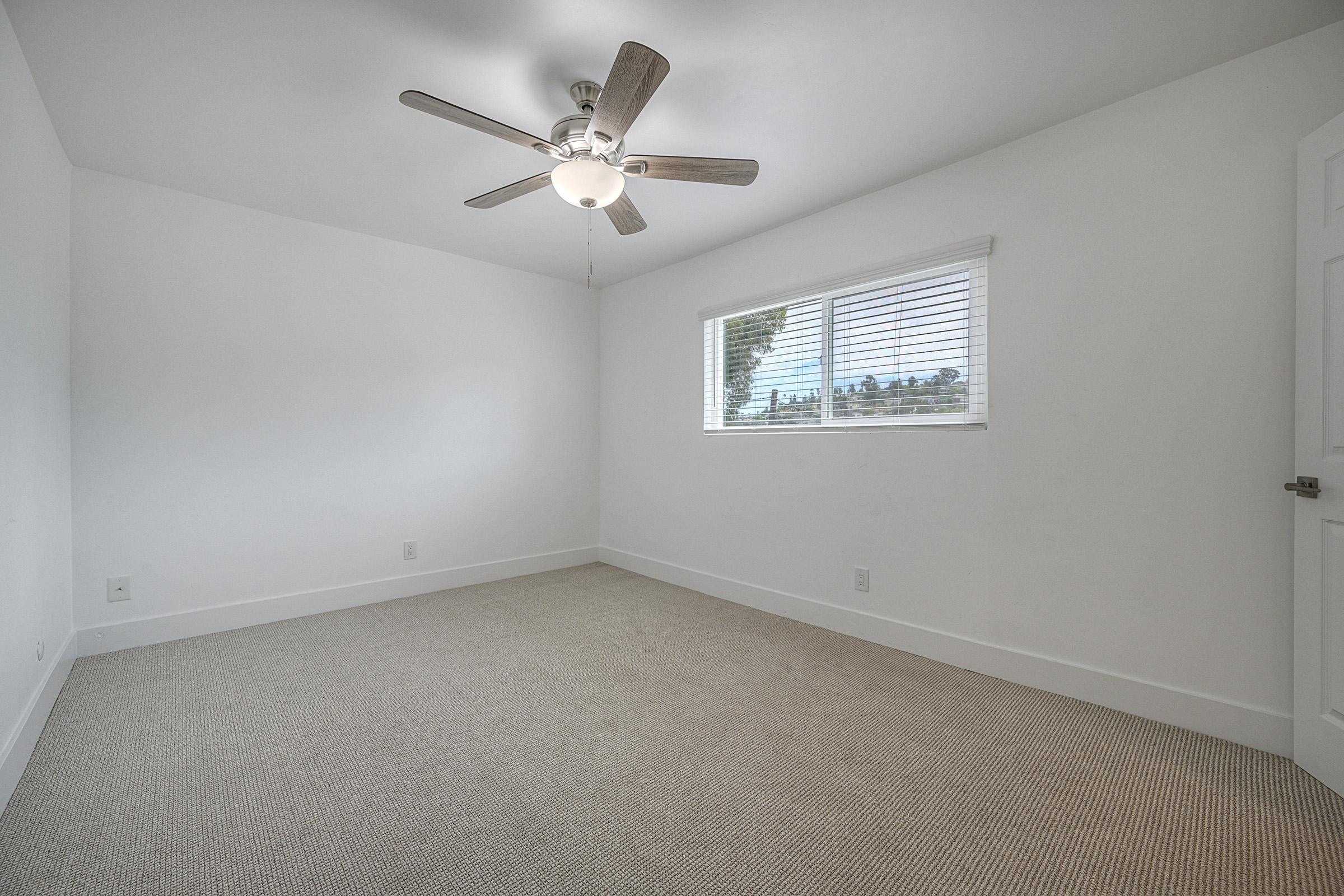 A clean, empty room with light-colored walls and a ceiling fan. Natural light enters through a window with blinds, illuminating the carpeted floor. The room has a simple design, making it suitable for various purposes.