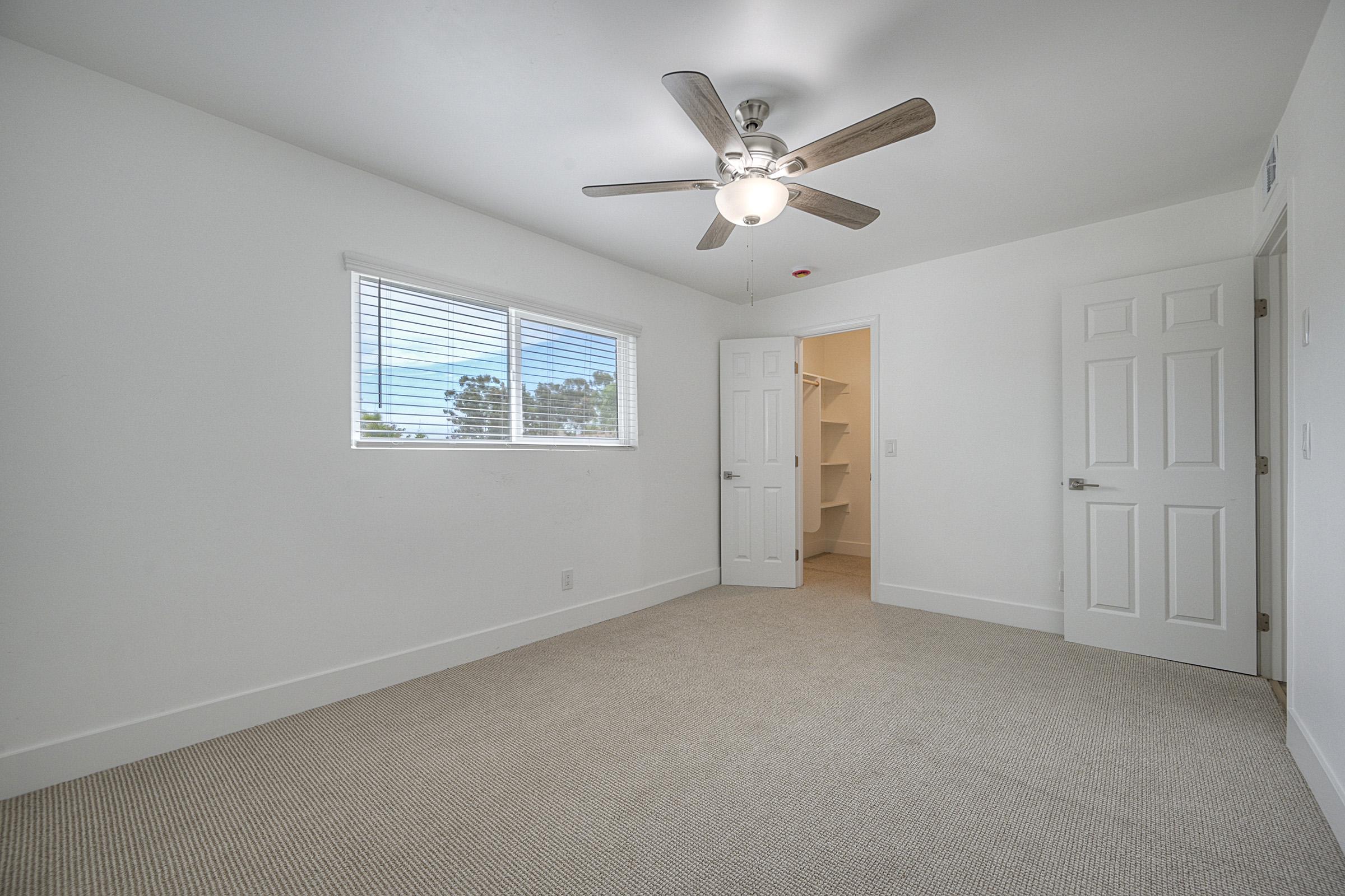 A bright, empty bedroom featuring light-colored walls and a ceiling fan. A window allows natural light to enter, and there are two closed doors, one leading to a closet or storage space. The floor is covered in light carpet, contributing to the room's airy ambiance.