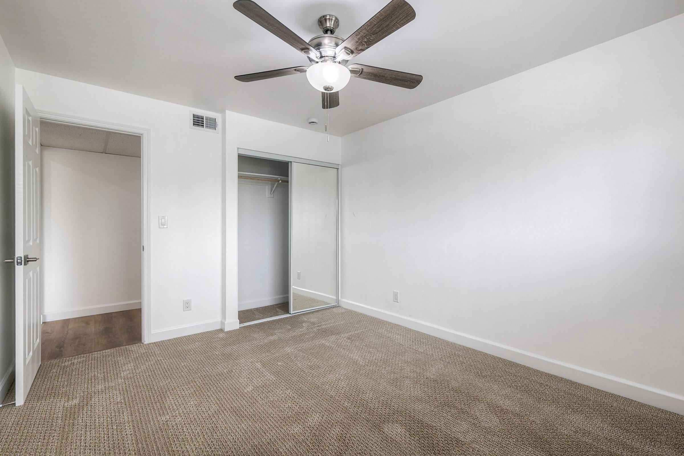 A bright, empty room featuring a light fan ceiling fixture, beige carpeting, and white walls. There's a mirrored closet on one side and an open door leading to another space. The room has a clean, minimalist aesthetic, ideal for personalized decoration.