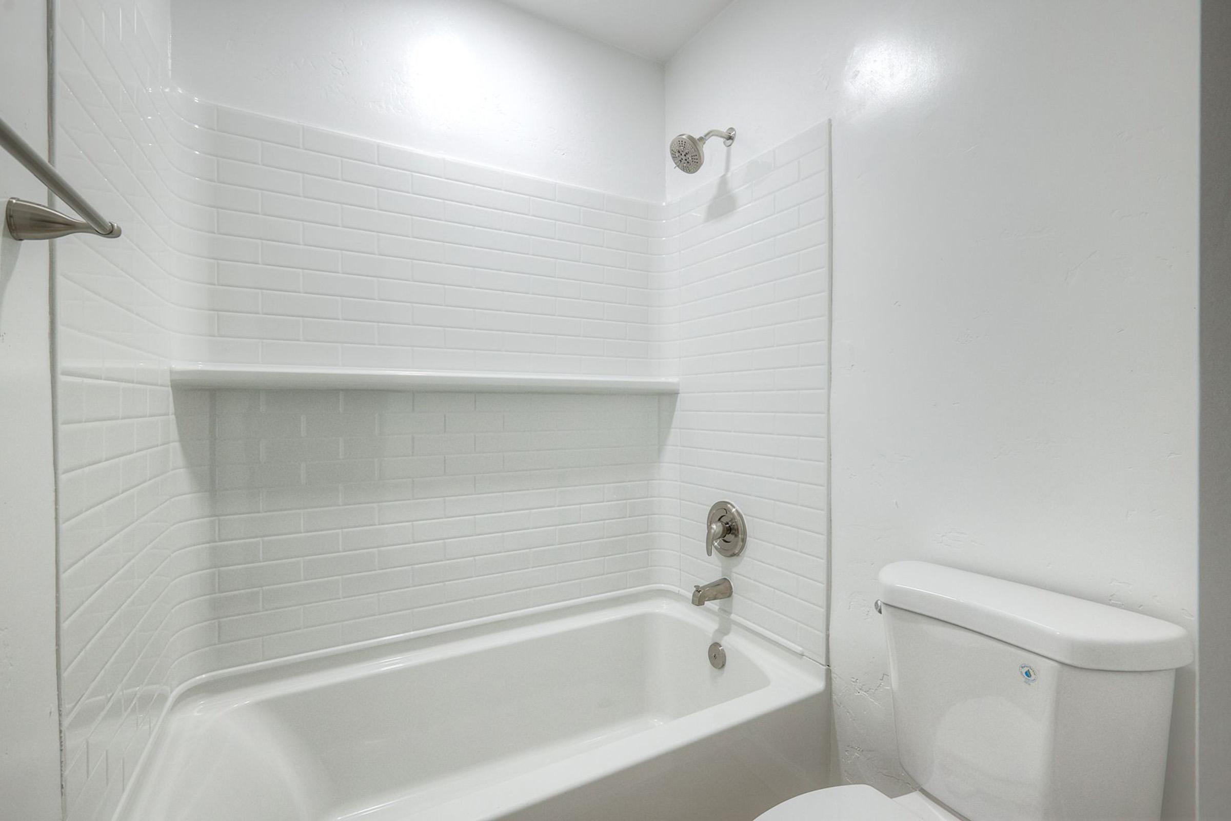 A clean and modern bathroom featuring a white bathtub and a shower with a silver faucet. The walls are lined with white subway tiles, and there are two shelves for storage. A white toilet sits beside the bathtub, all set against a bright, well-lit background.
