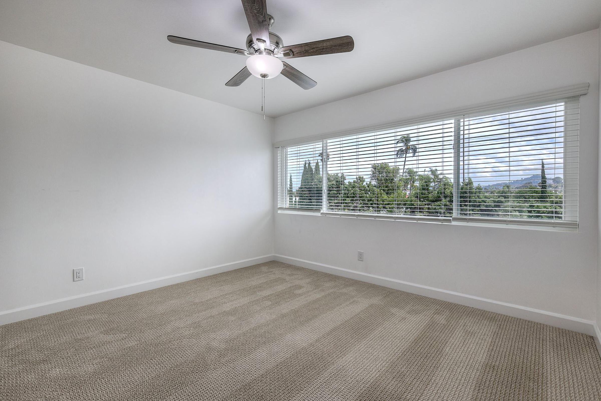 A spacious room with light-colored walls and a ceiling fan. There are large windows covered with blinds, offering a view of greenery outside. The floor is carpeted in a neutral tone, and the room is empty, providing a bright and airy atmosphere.