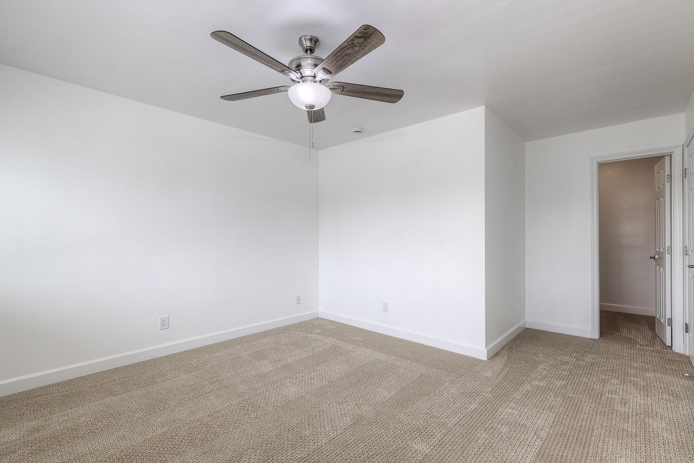 Spacious empty room featuring a ceiling fan, light-colored walls, and beige carpeting. A door on the right leads to another area, and the room is well-lit with natural light. Ideal for various uses such as a bedroom or office.