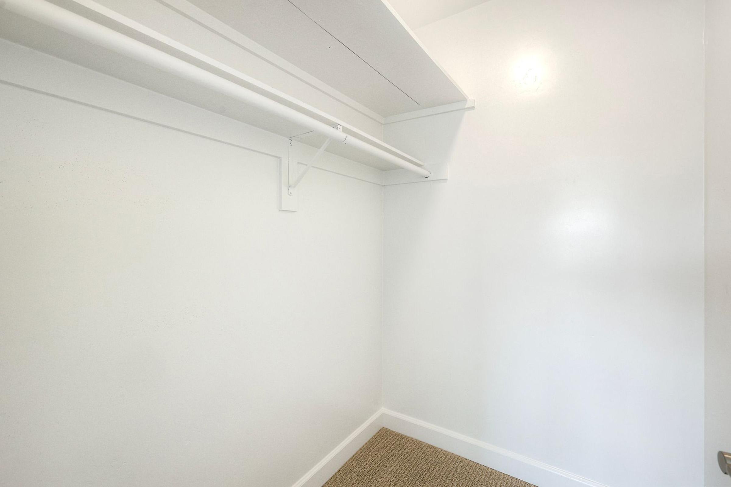 Empty closet with white walls and a single hanging rod. The floor is carpeted in a neutral color, and there's a small shelf above the rod. The space is well-lit and clean, creating a minimalist appearance.