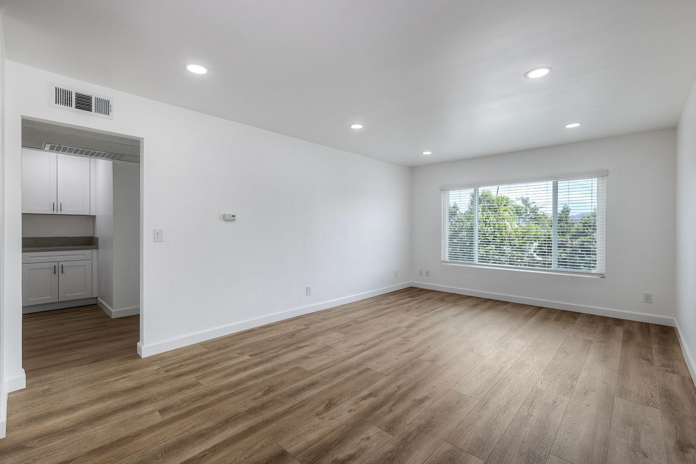 A spacious, bright living room featuring white walls and wooden flooring. Large window letting in natural light with views of greenery outside. In the background, a doorway leads to a kitchen area with white cabinets. The room is unfurnished, creating an open, inviting atmosphere.