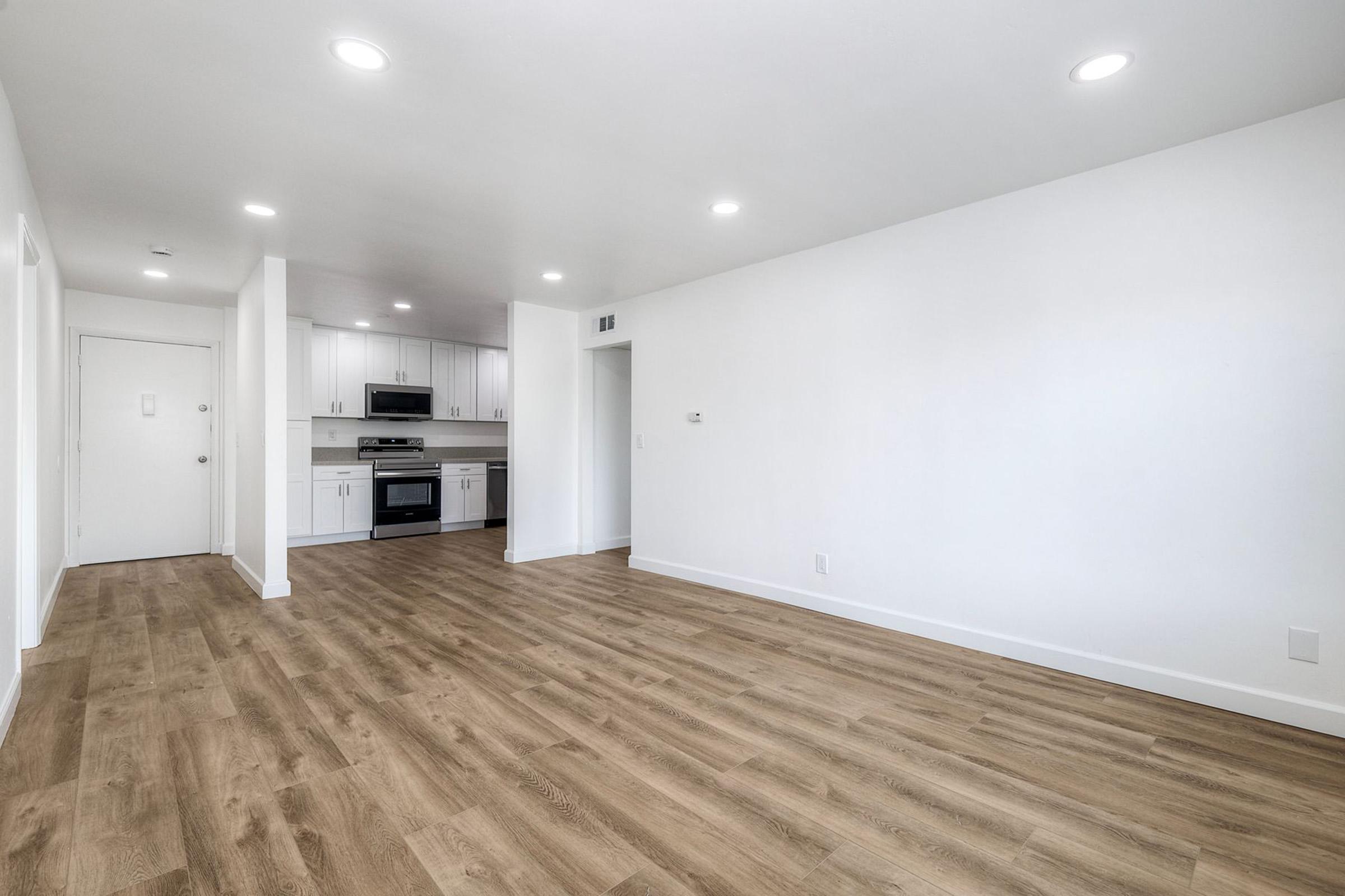A spacious, modern living area with light hardwood flooring. The room features white walls and ceiling lights, leading into a kitchen area with stainless steel appliances, including a stove and refrigerator, set against white cabinetry. The overall ambiance is bright and inviting.