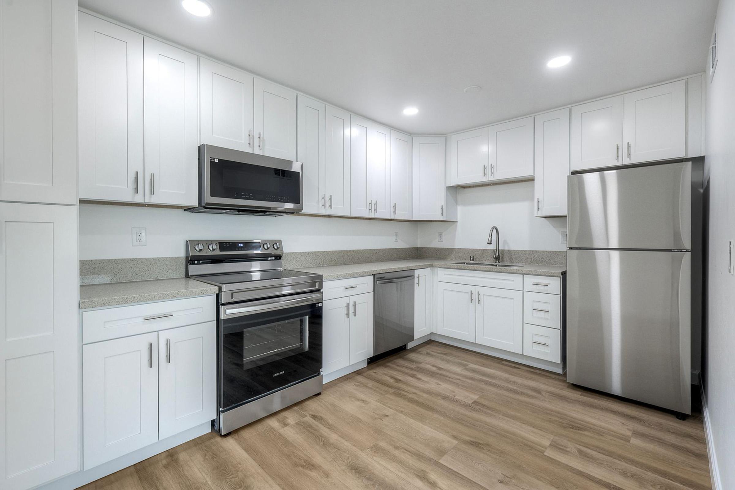 Modern kitchen featuring white cabinetry, stainless steel appliances including a microwave, oven, dishwasher, and refrigerator. The countertop is a light gray with a subtle pattern. The room has recessed lighting and a wood laminate floor, creating a clean and contemporary look.