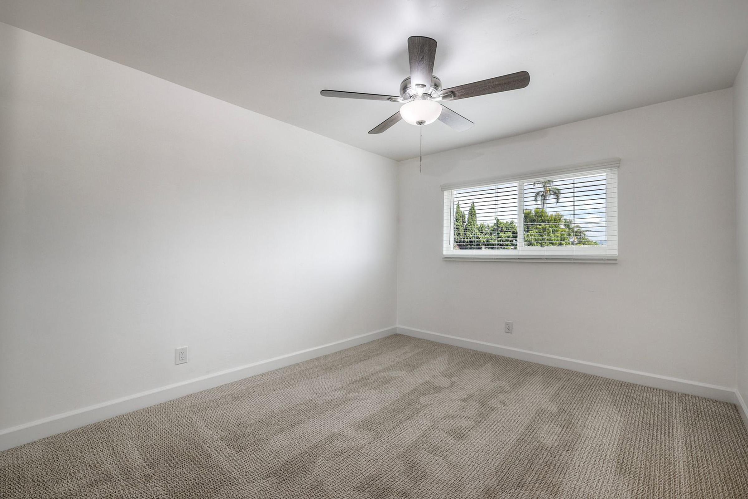 A clean, empty room featuring light-colored walls, carpet flooring, and a ceiling fan. A window with blinds allows natural light to enter, showcasing a view of greenery outside. The space is uncluttered, providing a blank canvas for decoration and furnishing.