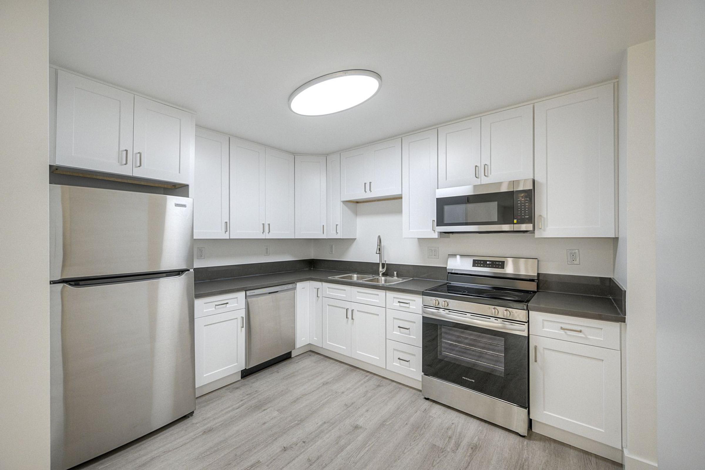 Modern kitchen featuring white cabinetry, stainless steel appliances including a refrigerator, microwave, and stove. The countertop is dark, contrasting with the light-colored cabinets. A round ceiling light illuminates the space, which has a clean and contemporary design with a wooden floor.