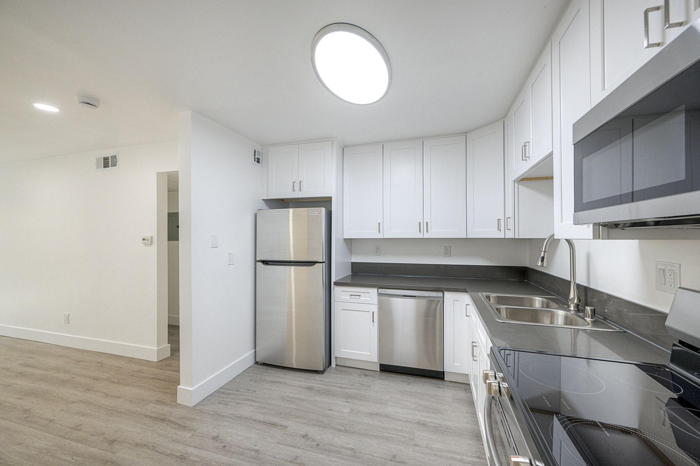 Modern kitchen with white cabinets, stainless steel appliances including a refrigerator, dishwasher, and microwave. The countertop is dark gray, and there is a sleek stove. The space features light-colored wood-style flooring and ample overhead lighting. The walls are painted white, creating a bright and airy atmosphere.