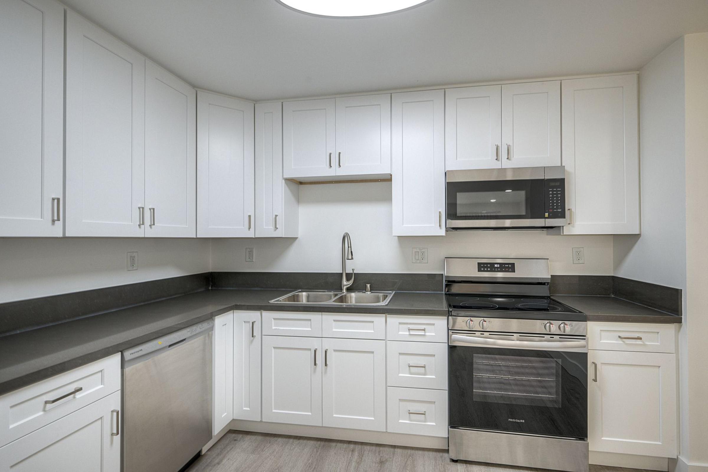 Modern kitchen featuring white cabinetry, a stainless steel microwave above an electric stove, and a double sink. The countertops are dark gray, complementing the light wood flooring. Appliances include a refrigerator and dishwasher, all designed for a clean and contemporary look.