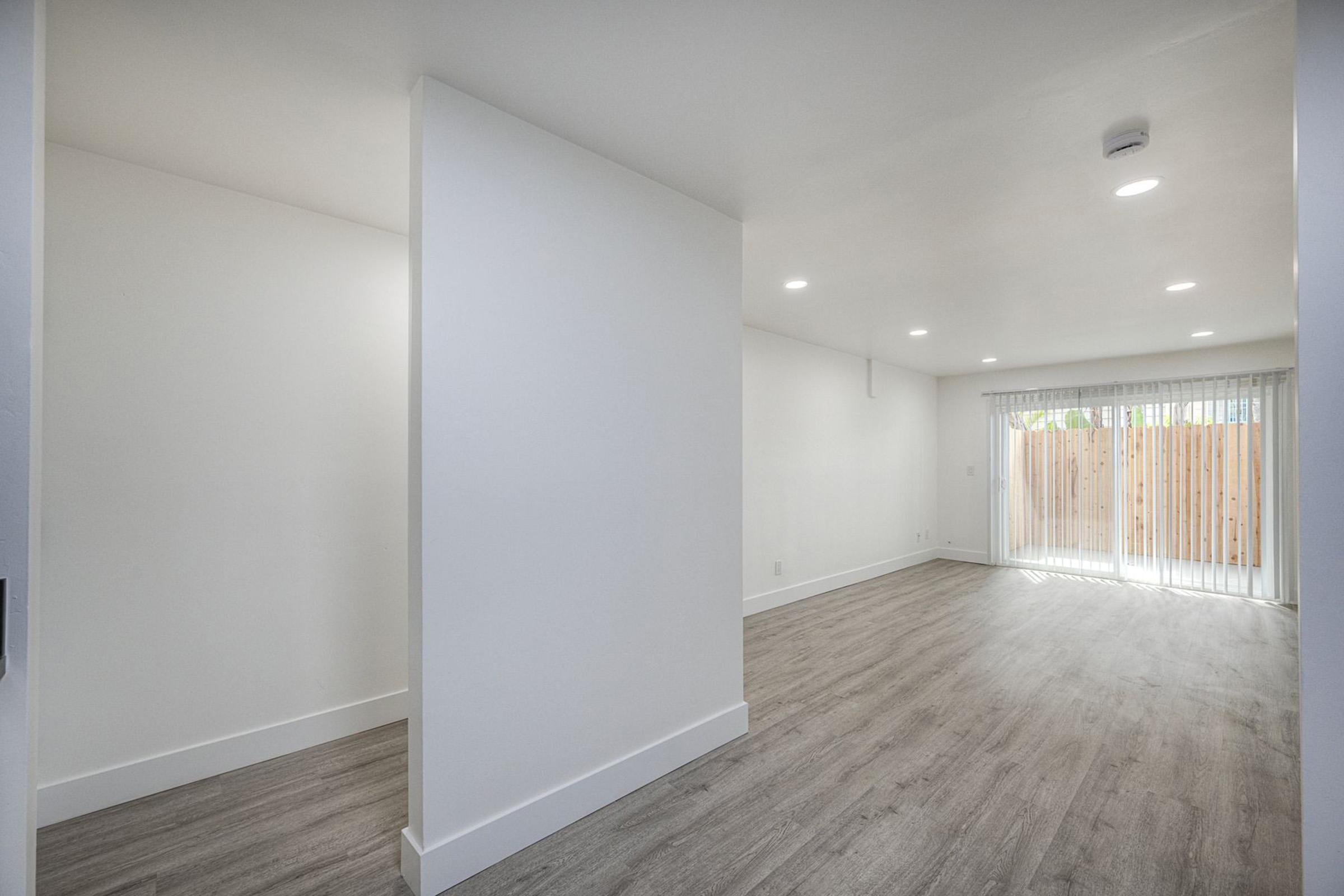 A bright and modern interior space featuring light-colored walls and a smooth, wooden floor. A partially divided area on the left leads to an open living space with a large window covered by sheer curtains, allowing natural light to fill the room.