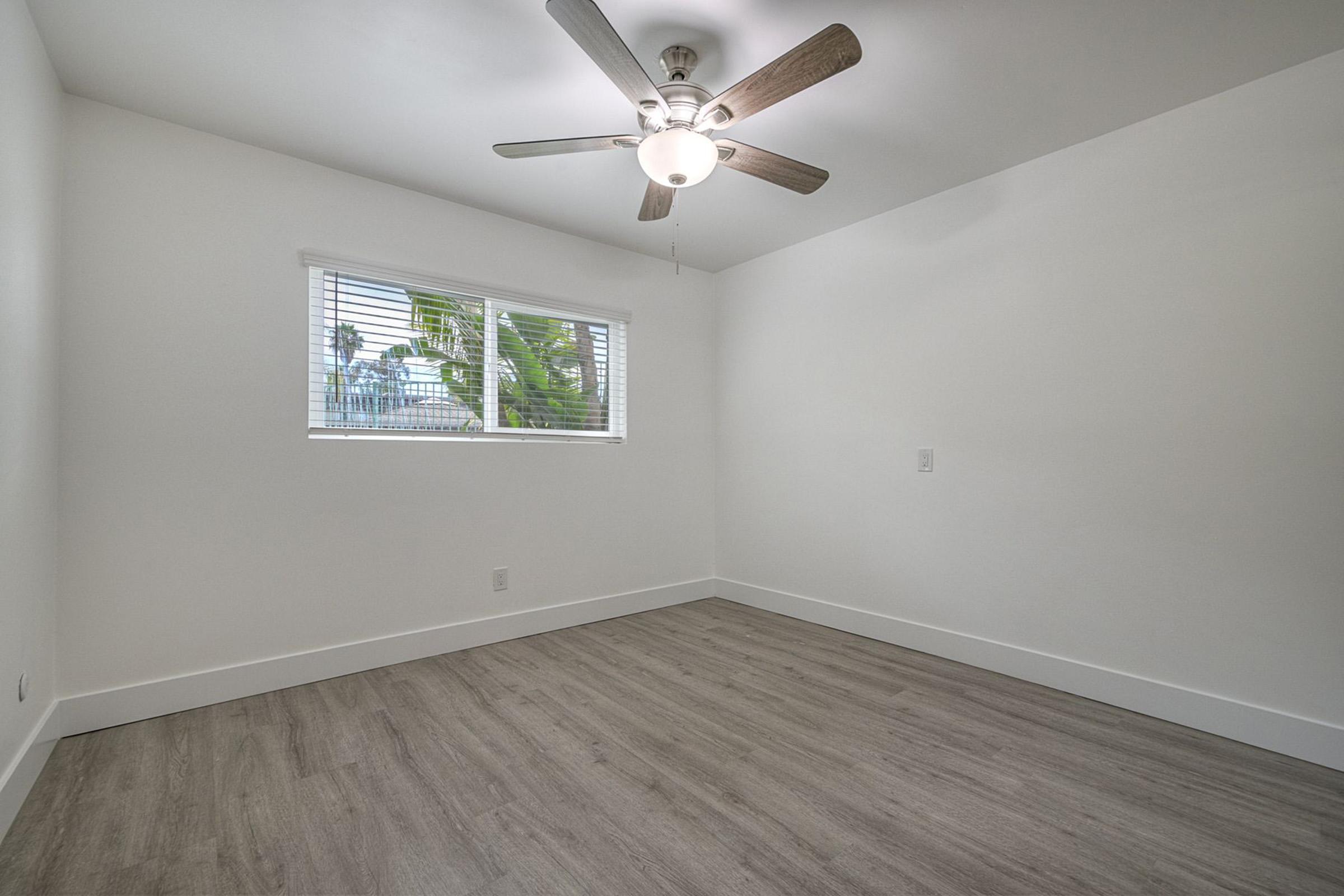 A bright, minimalist bedroom with light-colored walls and wood-like flooring. A ceiling fan is mounted in the center, and there’s a window with blinds allowing natural light to enter. The room appears clean and uncluttered, ideal for various decorating styles.