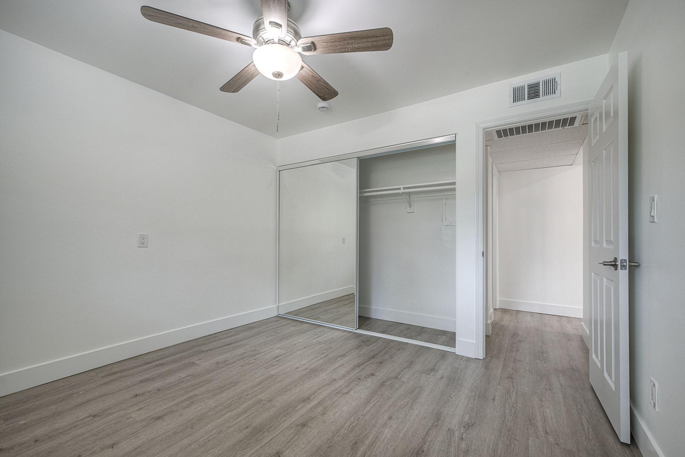A bright, empty room featuring a ceiling fan and mirrored closet doors. The walls are white, and the floor has light-colored, wood-like laminate. A door is visible on the right, leading to another space, while the overall design conveys a modern and spacious feel.