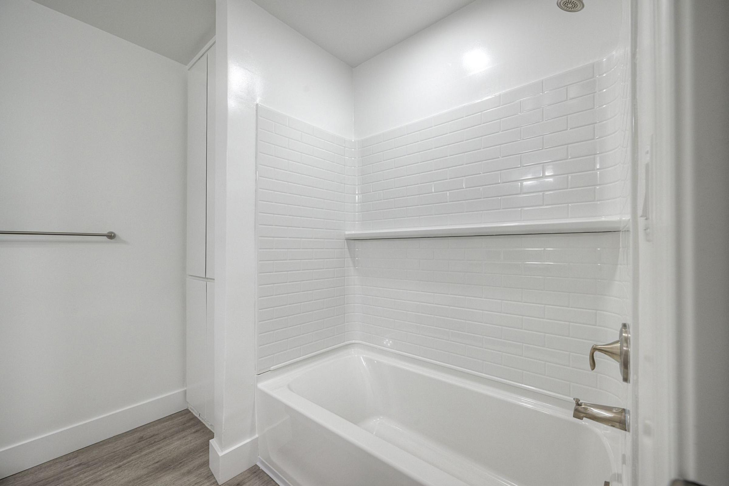 A clean, modern bathroom featuring a white bathtub with a tiled wall in the background. The walls are painted white, and there is a sleek grab bar on the left. The flooring is light wood, creating a bright and minimalist aesthetic.