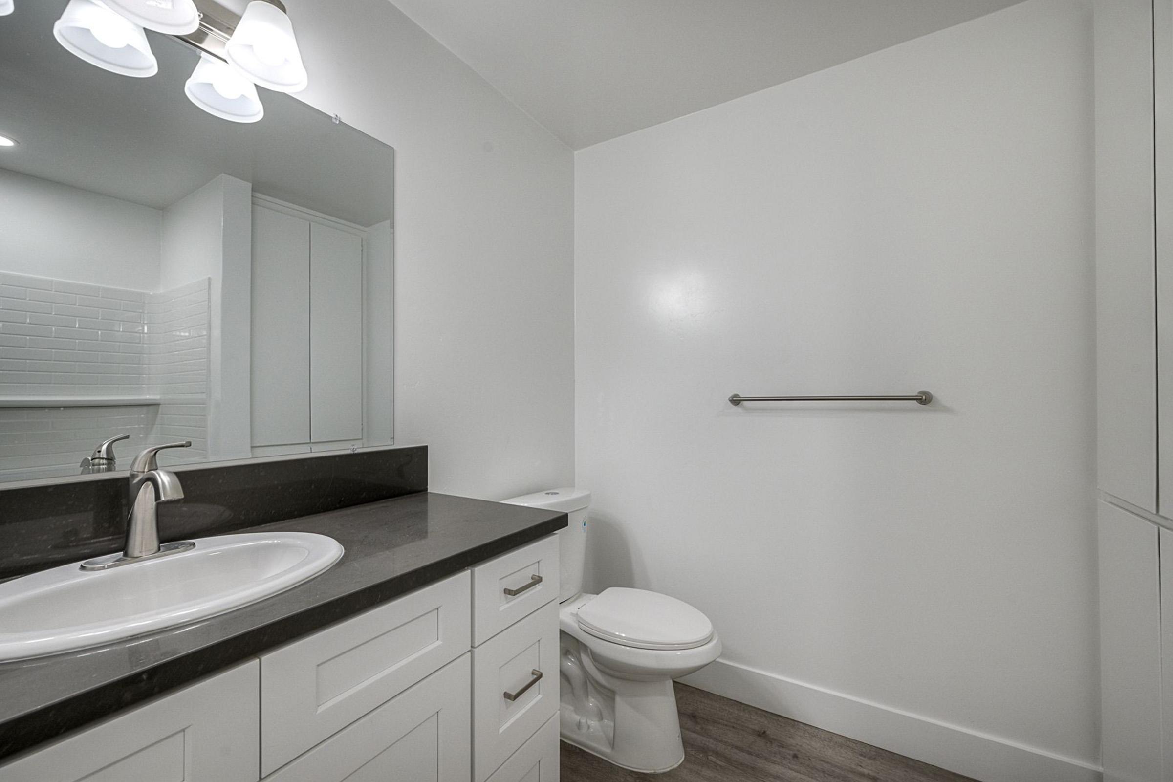 Modern bathroom featuring a white vanity with a round sink and a dark countertop. A large mirror is mounted above the sink, with three light fixtures above it. The walls are painted white, and there is a toilet and a towel bar. The floor has light wood-like tiles.