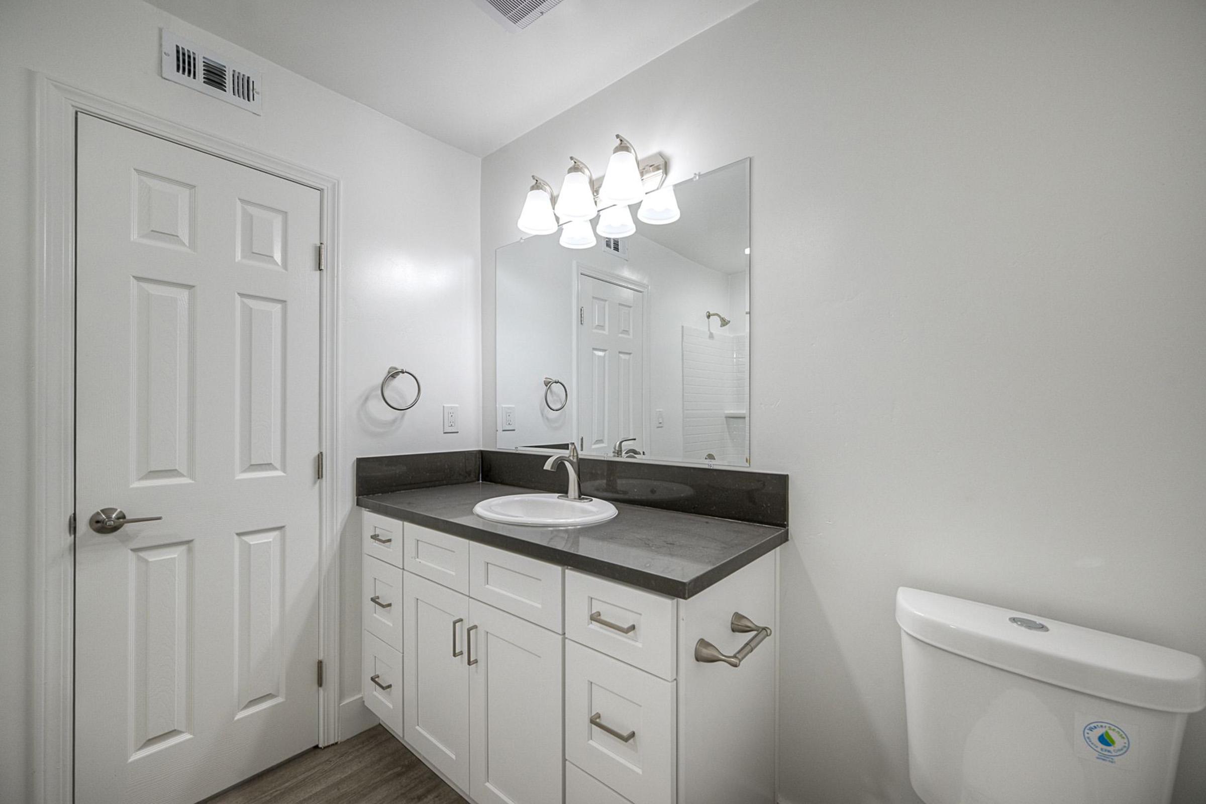 A clean, modern bathroom featuring a white vanity with a sink, gray countertop, and mirror. There's a white toilet on the right, a door leading to another room, and simple wall fixtures. The space is well-lit with overhead lights, and the walls are painted a light color, creating a bright atmosphere.