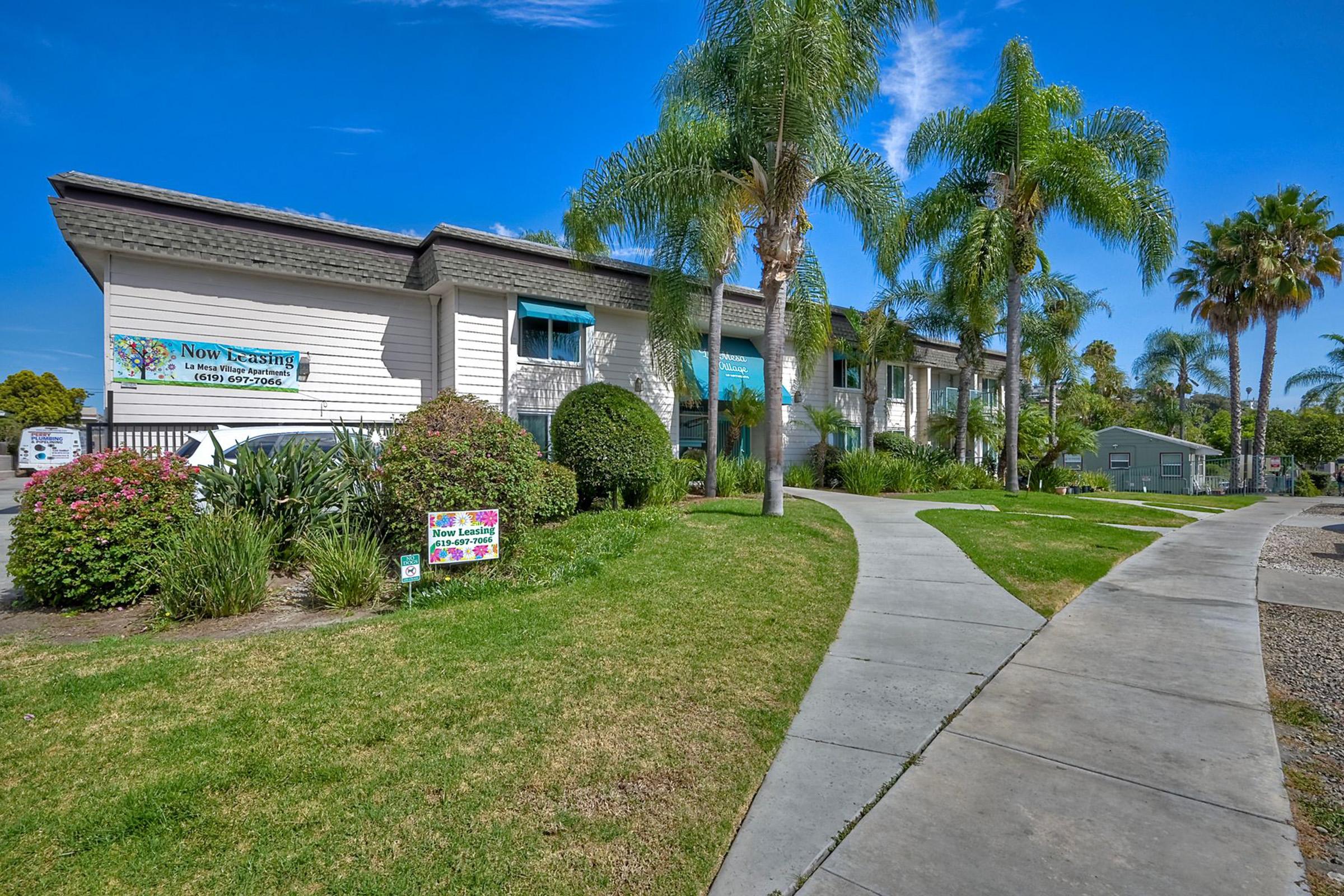 A walkway leading to a light-colored building with palm trees and green landscaping. A sign on the building mentions "Now Leasing" and there are colorful plants in the foreground. The sky is clear and blue, creating a bright and inviting atmosphere.