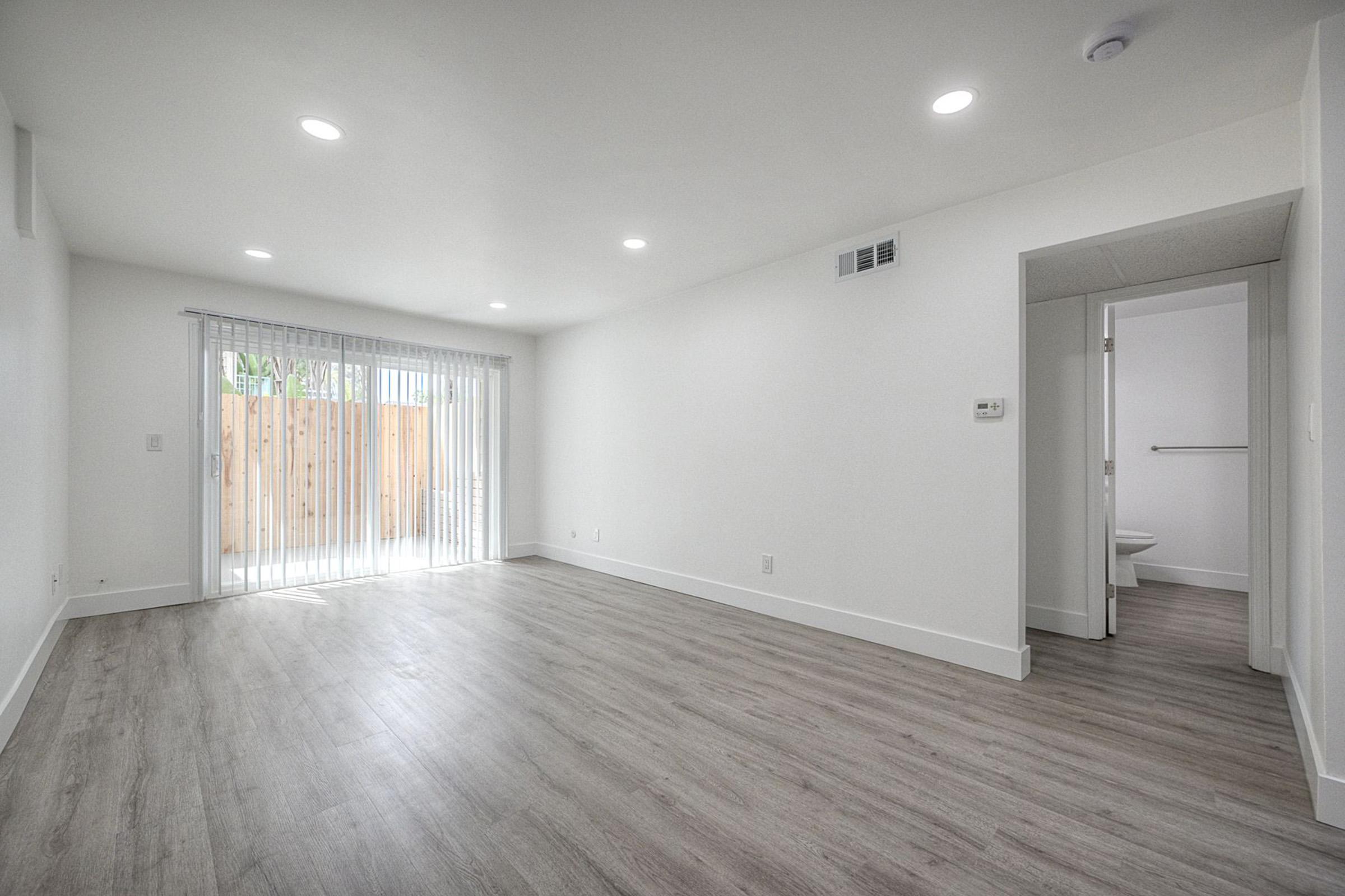 A spacious, empty room with light-colored walls and laminate flooring. Natural light streams in through a sliding glass door leading to a small outdoor space. There's a doorway to a bathroom on the right, and the overall design is modern and minimalistic.