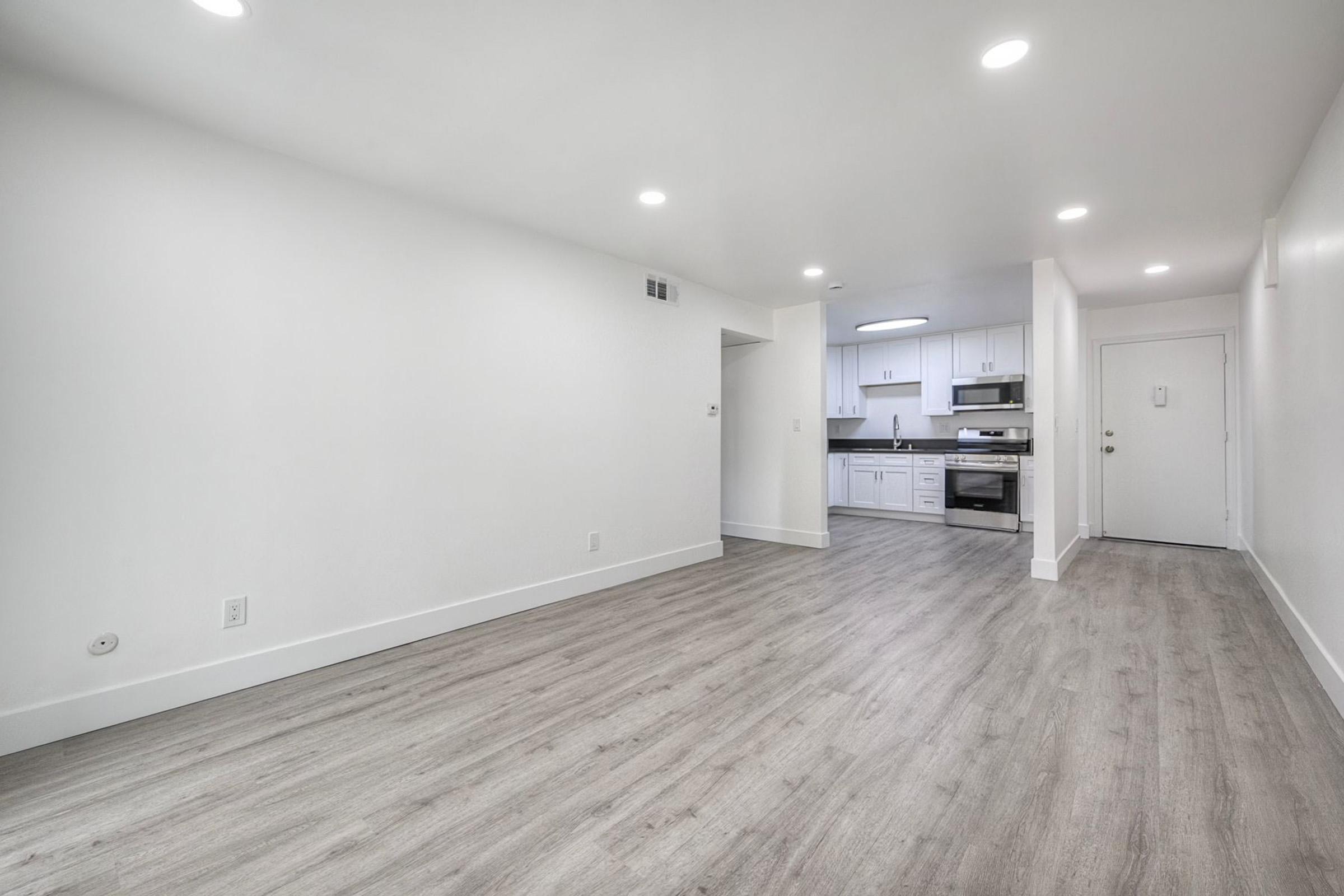 A spacious, modern living area with light gray flooring and white walls. To the left, there's a doorway leading to the kitchen, which features sleek white cabinetry and stainless steel appliances. The room is well-lit with recessed lighting and has an open layout, providing a bright and airy atmosphere.