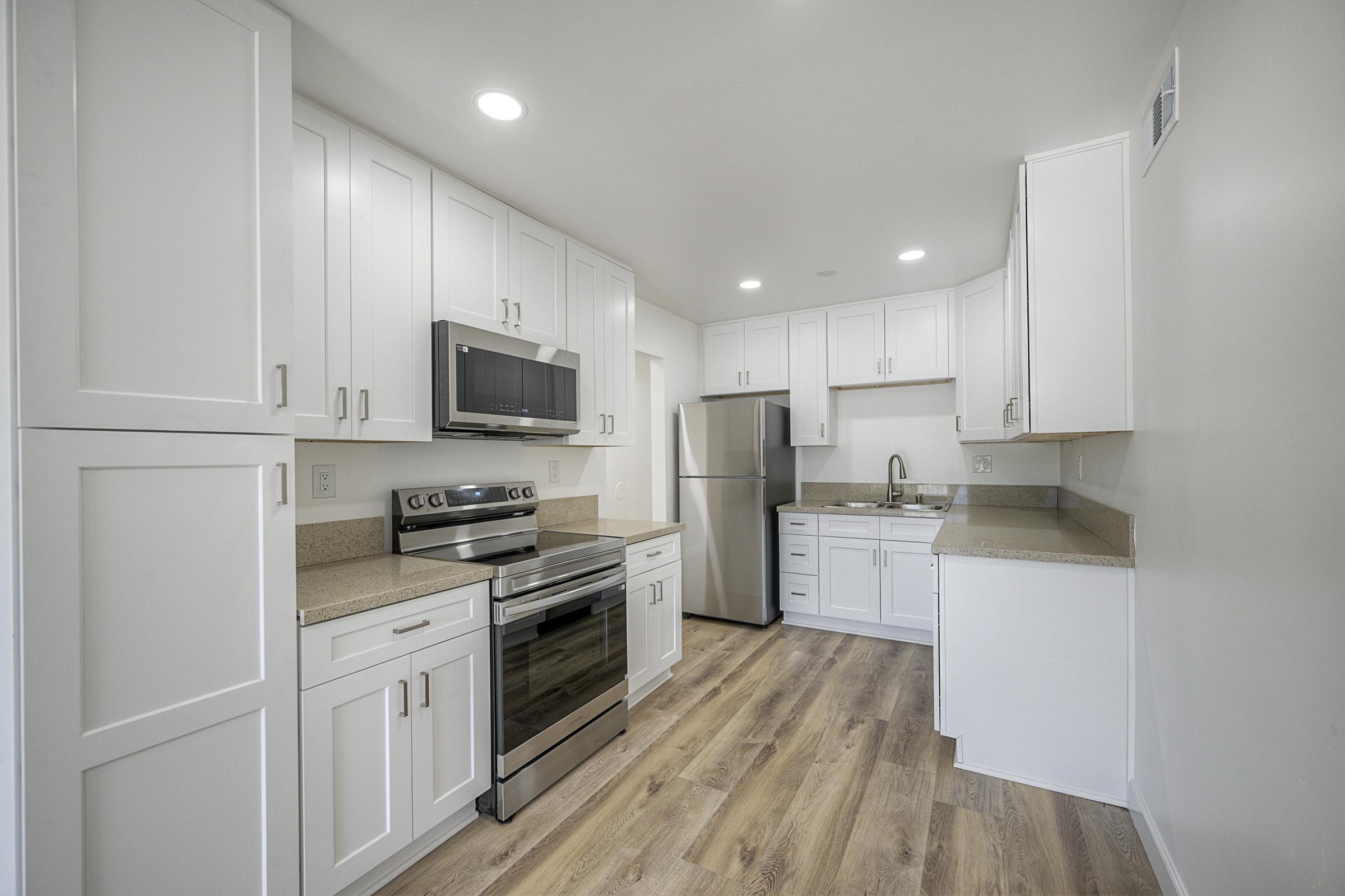 Modern kitchen featuring white cabinetry, stainless steel appliances including a microwave and oven, a sink with a faucet, and ample counter space. The floor is made of wood-like laminate, and there are bright overhead lights illuminating the area.
