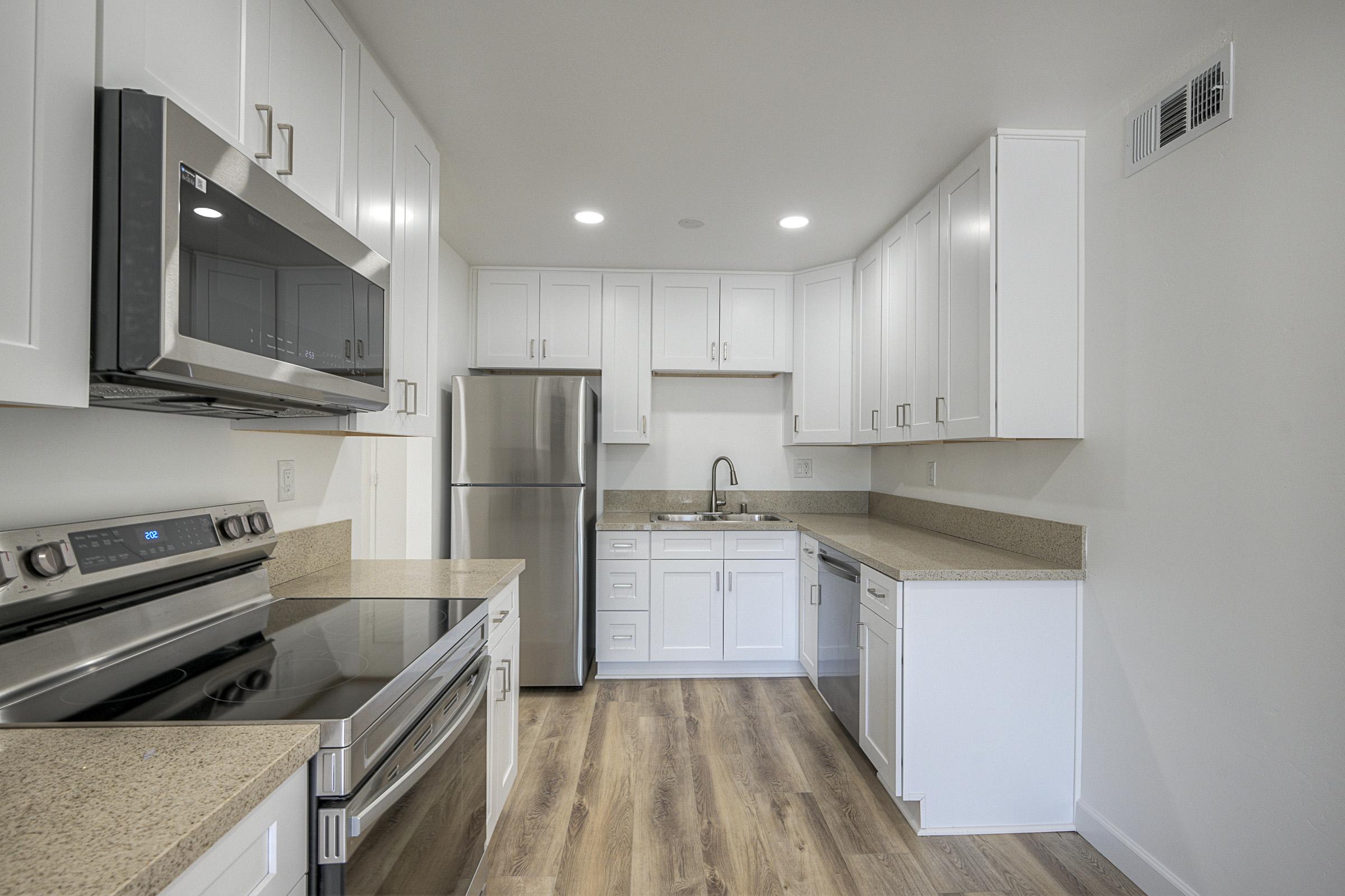 Modern kitchen featuring white cabinets, stainless steel appliances including a microwave and refrigerator, and a large stove with an oven. The countertops are a light-colored stone, and the flooring is a wood laminate. Bright lighting fixtures illuminate the space, creating an open and airy atmosphere.