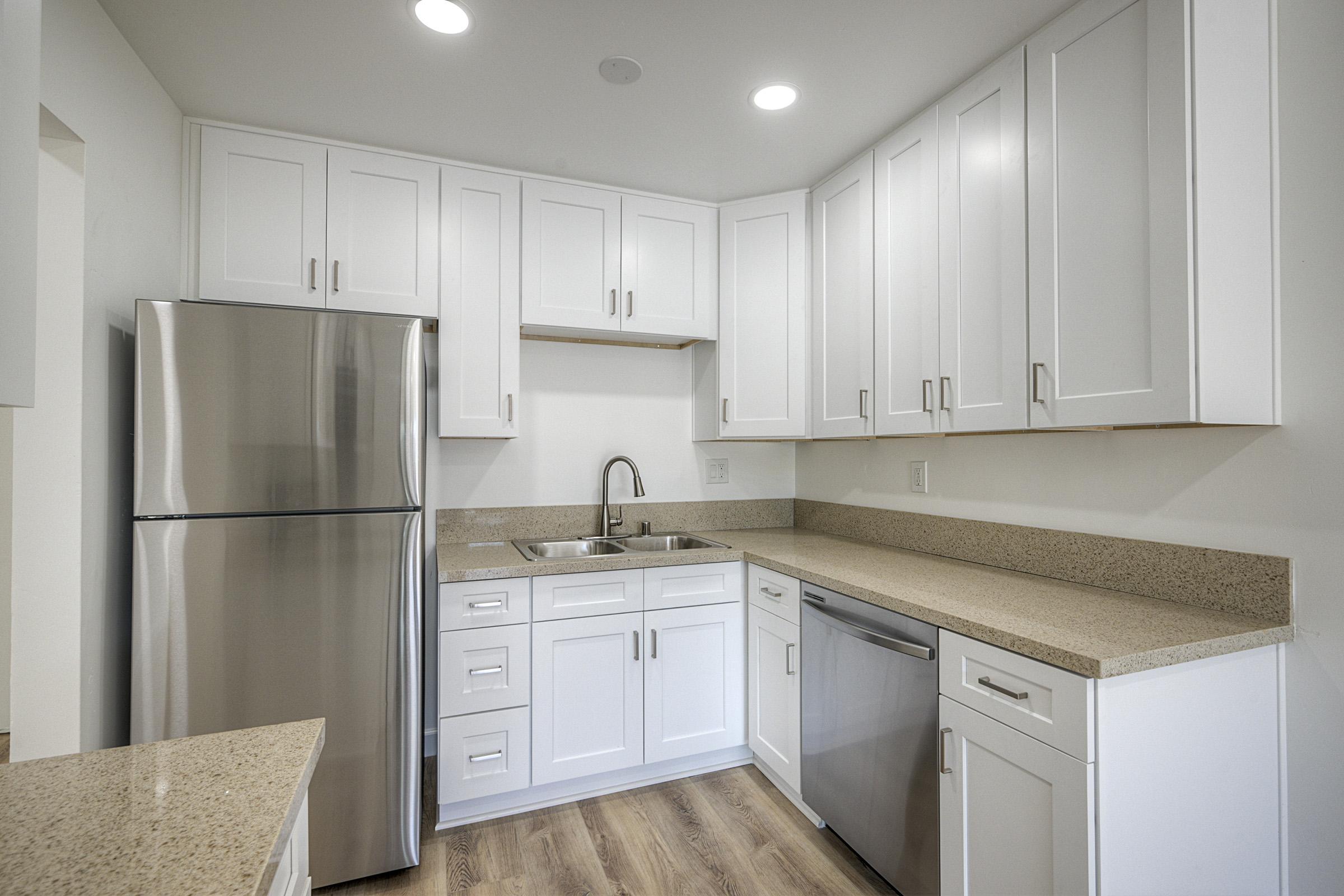Modern kitchen featuring white cabinets, a stainless steel refrigerator, a sink, and a dishwasher. The countertop is made of a light-colored stone, and the space is well-lit with overhead lights. The flooring is wood-like, adding warmth to the contemporary design.
