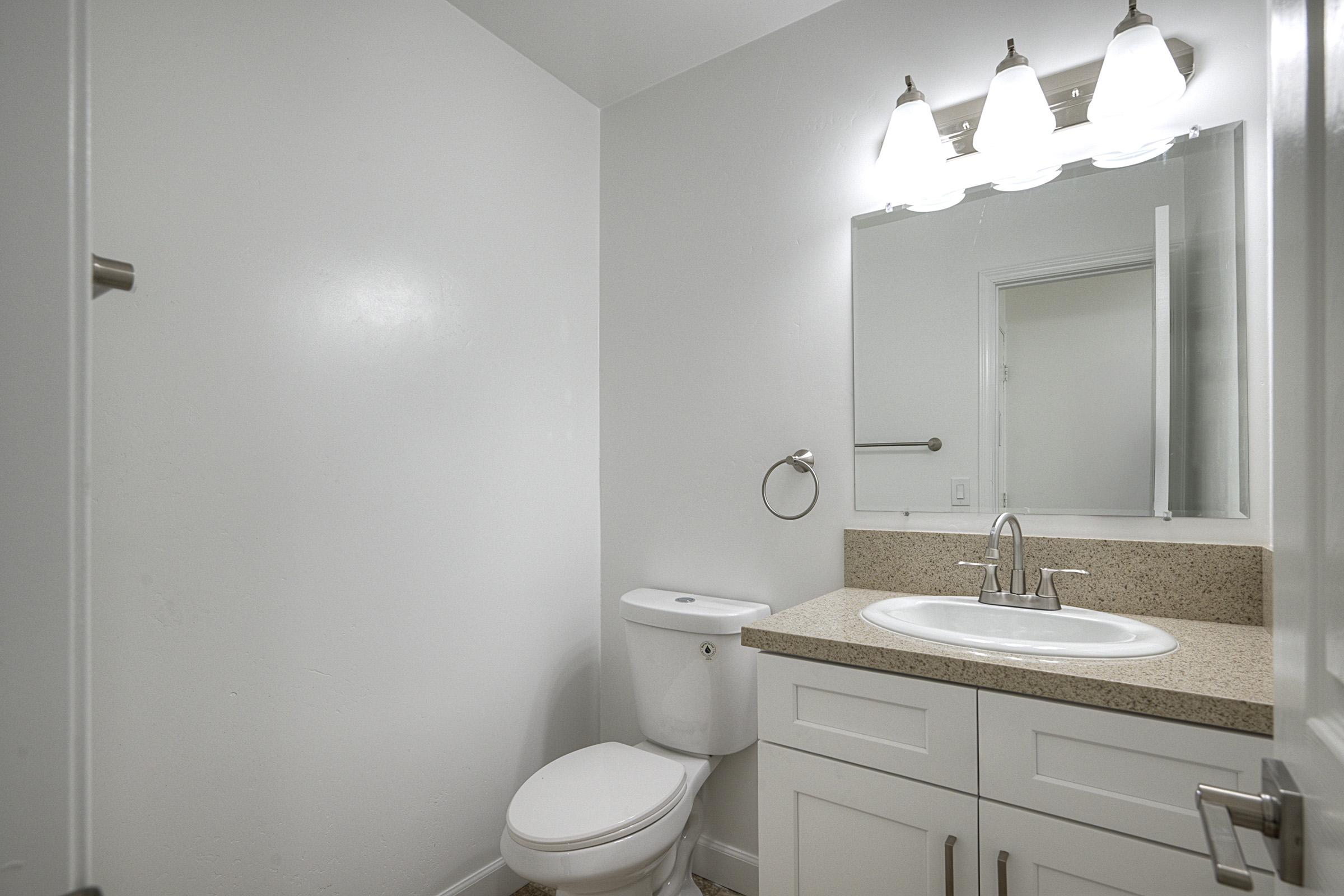 A clean, modern bathroom featuring a white cabinet with a sink and a countertop, a mirror above the sink, a toilet, and a light fixture with three bulbs. The walls are plain white, and the flooring is light-colored tiles. A towel ring is mounted on the wall near the sink.