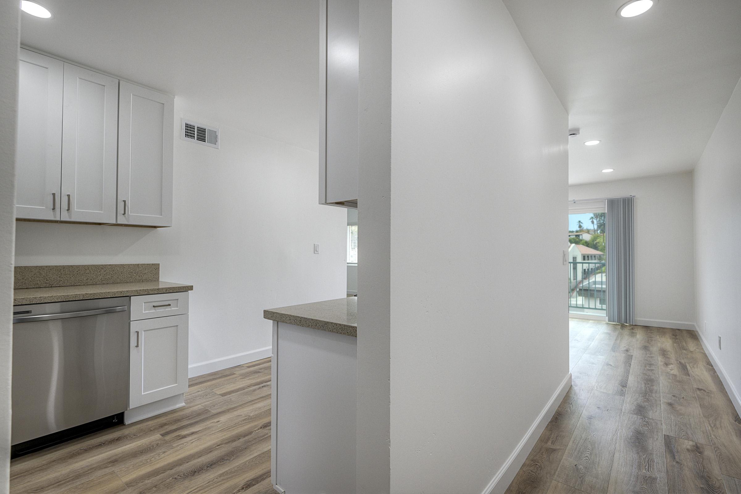 A modern kitchen area with white cabinetry and stainless steel appliances, transitioning to an open living space with large windows. The flooring is light wood, and the walls are painted white, creating a bright, airy atmosphere. A sliding door leads to a balcony with outdoor views.