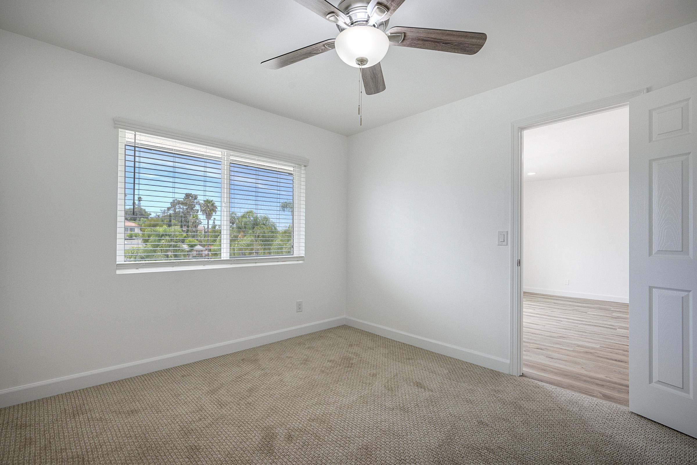 A bright, empty room with beige carpet and white walls. A ceiling fan is mounted above, and a window with white blinds allows natural light to enter. In the background, there's an open doorway leading to another light-filled room. The overall atmosphere is clean and minimalistic.