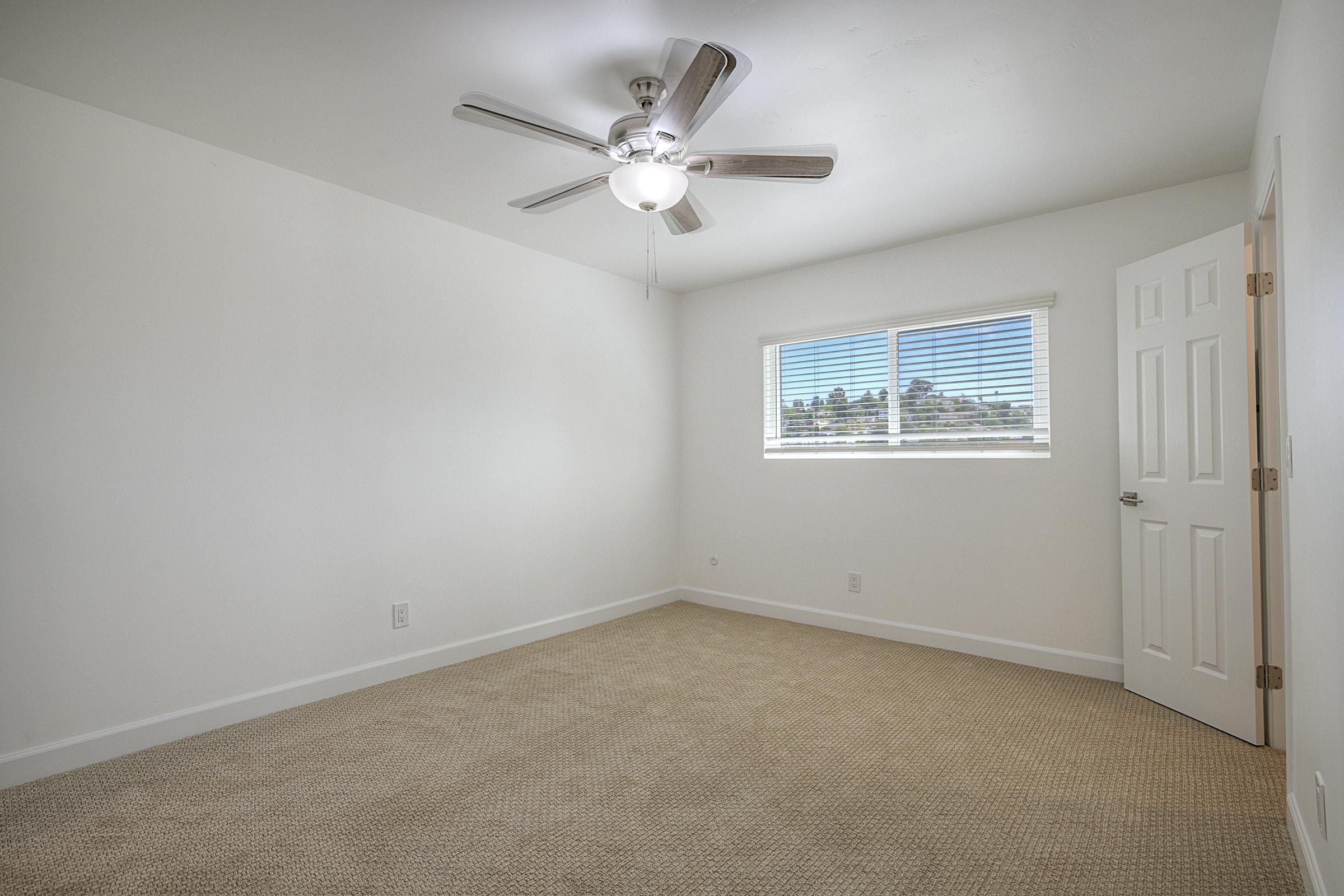 A clean, empty room with light-colored walls and a ceiling fan. It features a window with blinds, allowing natural light, and a beige carpeted floor. A door on the right leads to another space, suggesting the room is part of a larger home. The overall atmosphere is bright and minimalist.