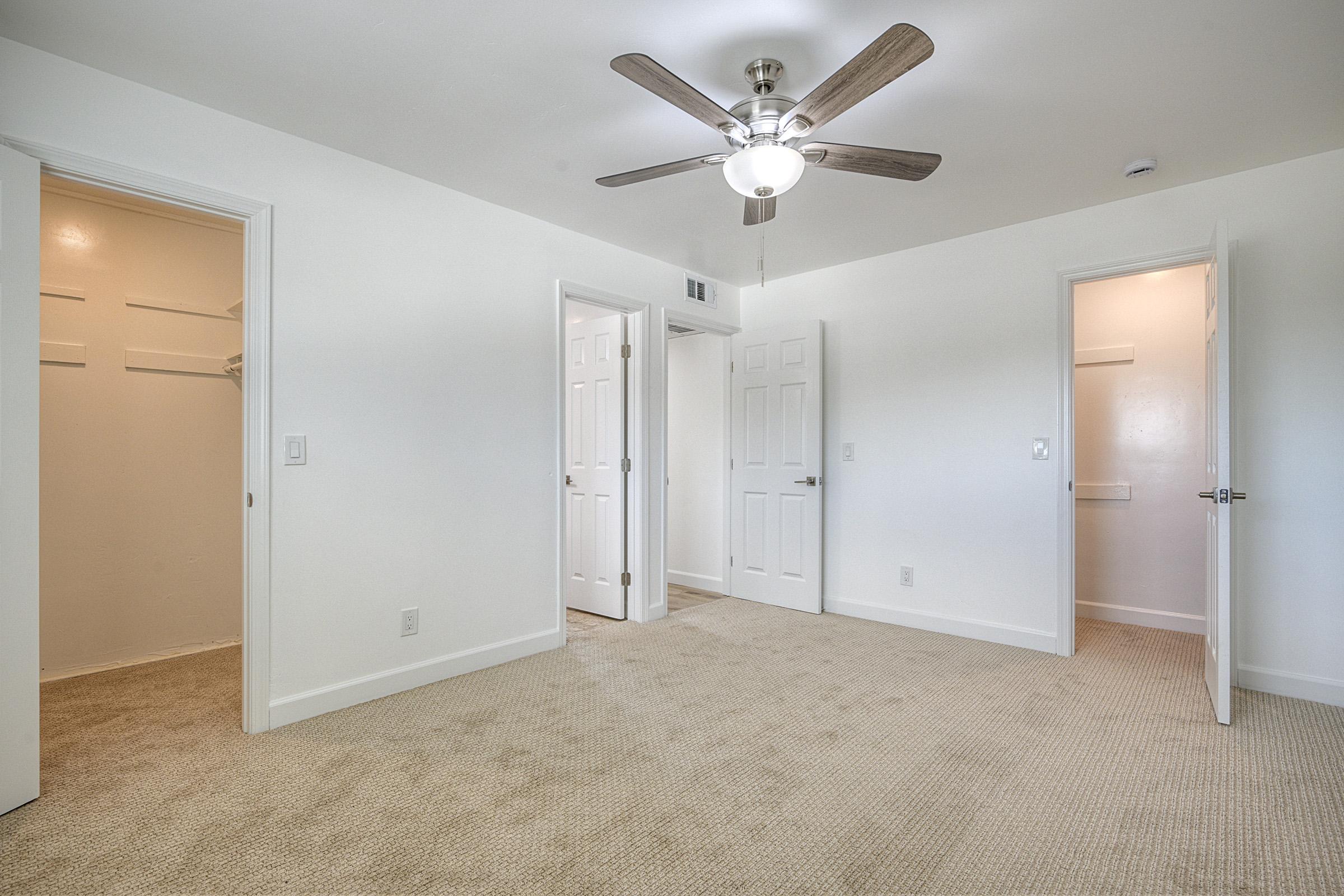 A spacious, empty room featuring light-colored walls and beige carpet. There are two doors leading to closets and one door leading outside the room. A ceiling fan is installed in the center, providing a modern touch to the minimalist decor. The ambient lighting creates a bright, inviting atmosphere.