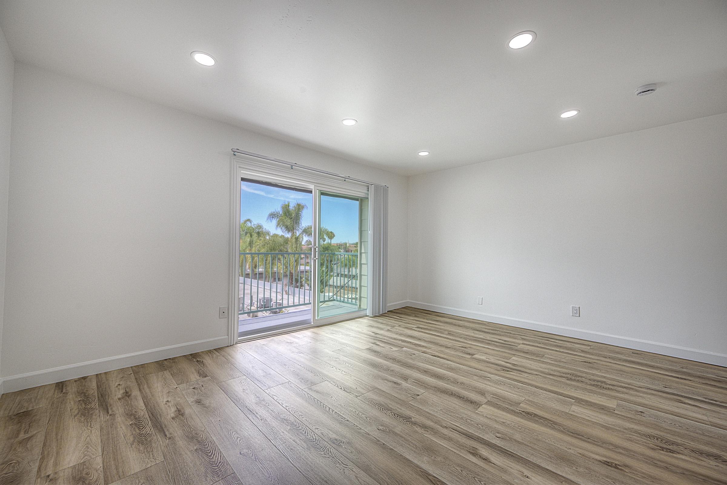 A spacious, empty room with light-colored walls and laminate flooring. A large sliding glass door leads to a balcony with a view of palm trees and blue sky. The room is well-lit with recessed lighting, creating a bright and airy atmosphere.